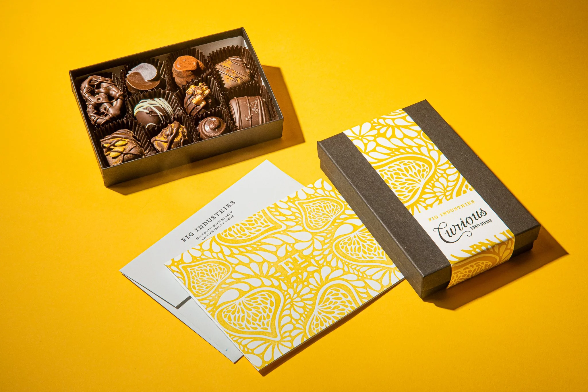 Image of mixed box of chocolates on yellow background with a letter pressed card next to it. Box is wrapped in a band featuring a fig illustrated pattern. The same fig pattern appears on the letter pressed card. Fig Industries Curious Confections.