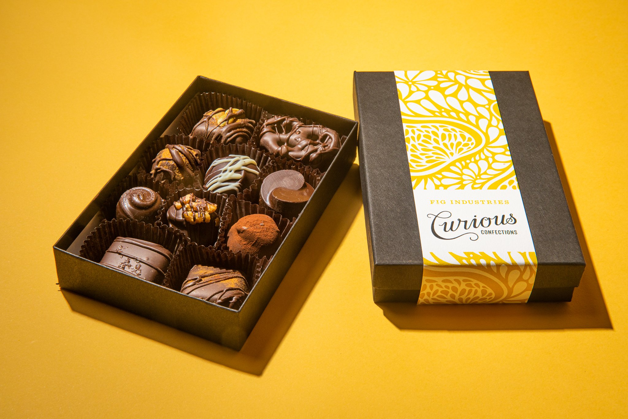Image of mixed box of chocolates on yellow background. Box is wrapped in a band featuring an illustrated fig pattern. Fig Industries Curious Confections.