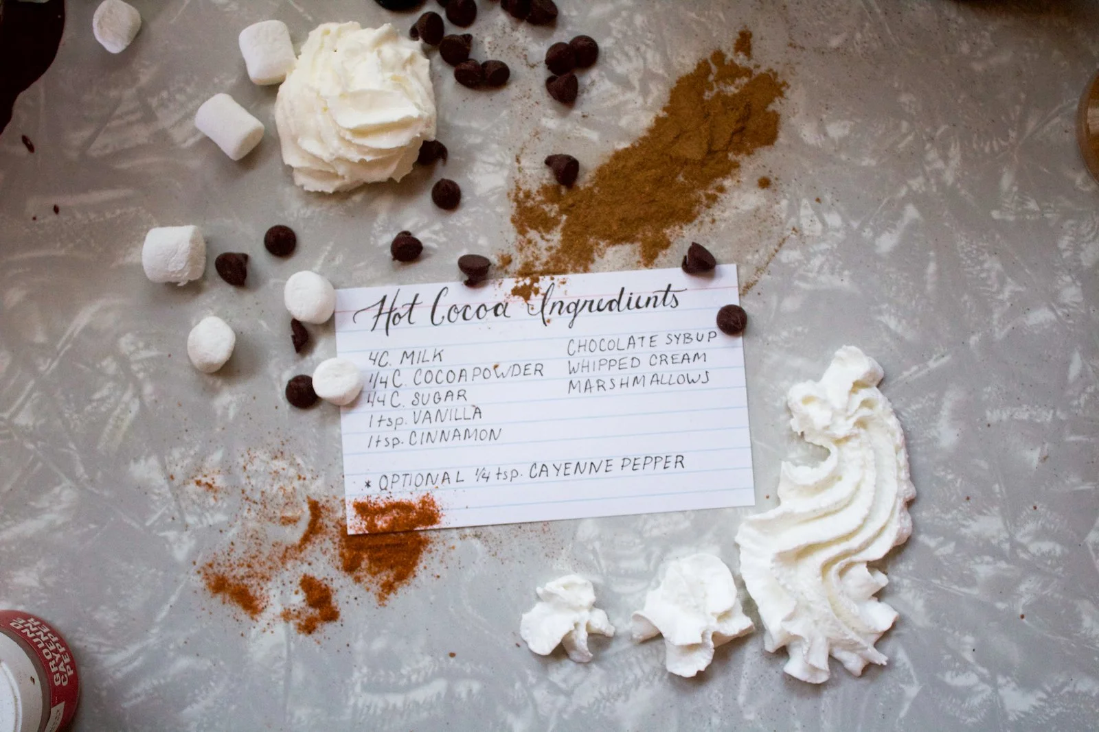 Image of hand written recipe for hot cocoa, surrounded by ingredients scattered around notecard.