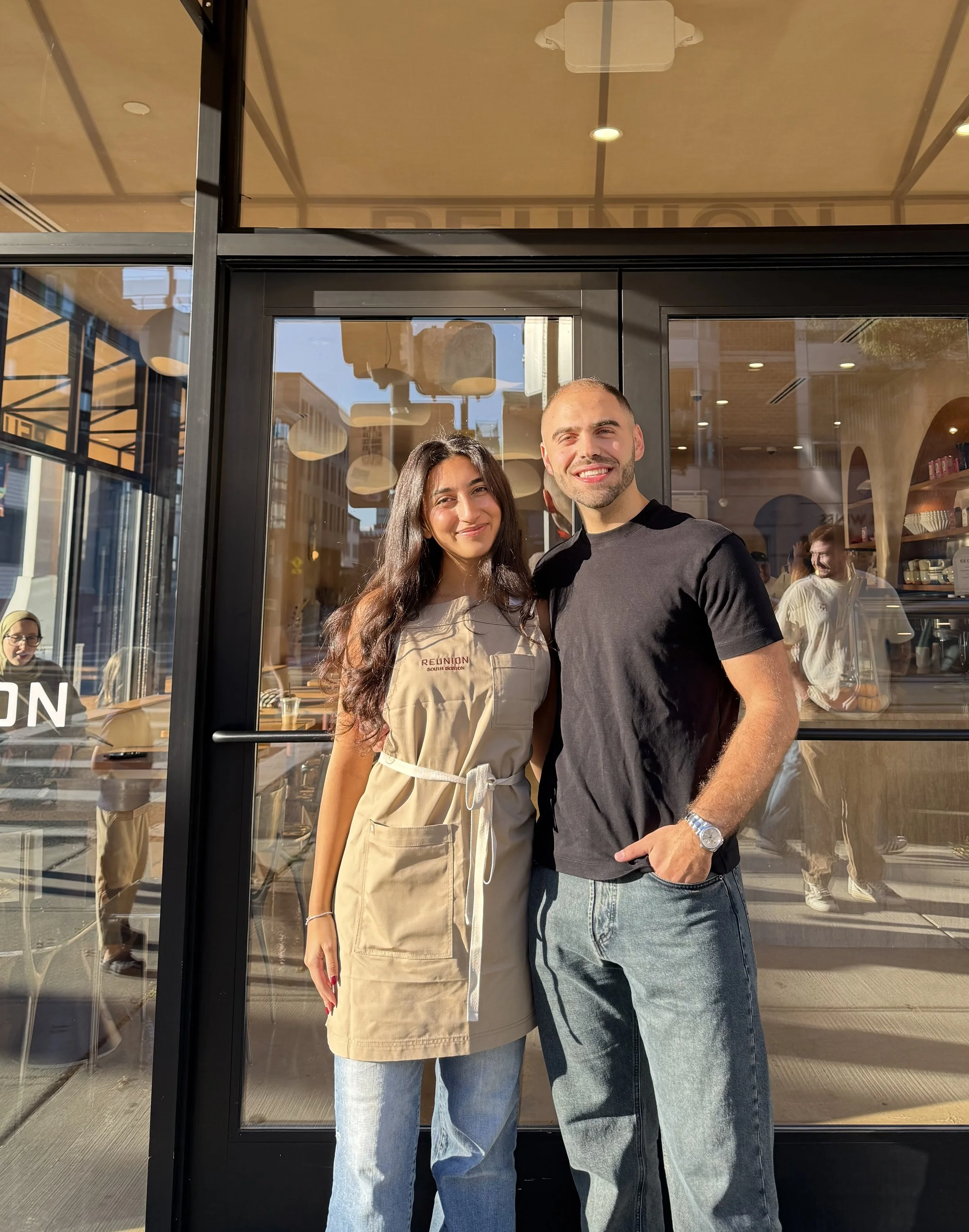 A woman and man standing outside a cafe, smiling at the camera, with the woman dressed in a beige apron and the man in a black t-shirt, sunlight shining on them.
