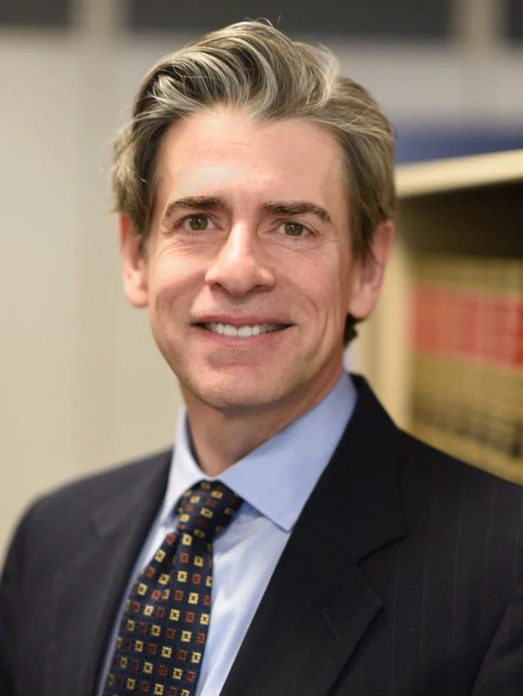 Professional headshot of a man with light brown hair, wearing a dark suit, light blue shirt, and a colorful patterned tie, standing in an office environment with bookshelves in the background.