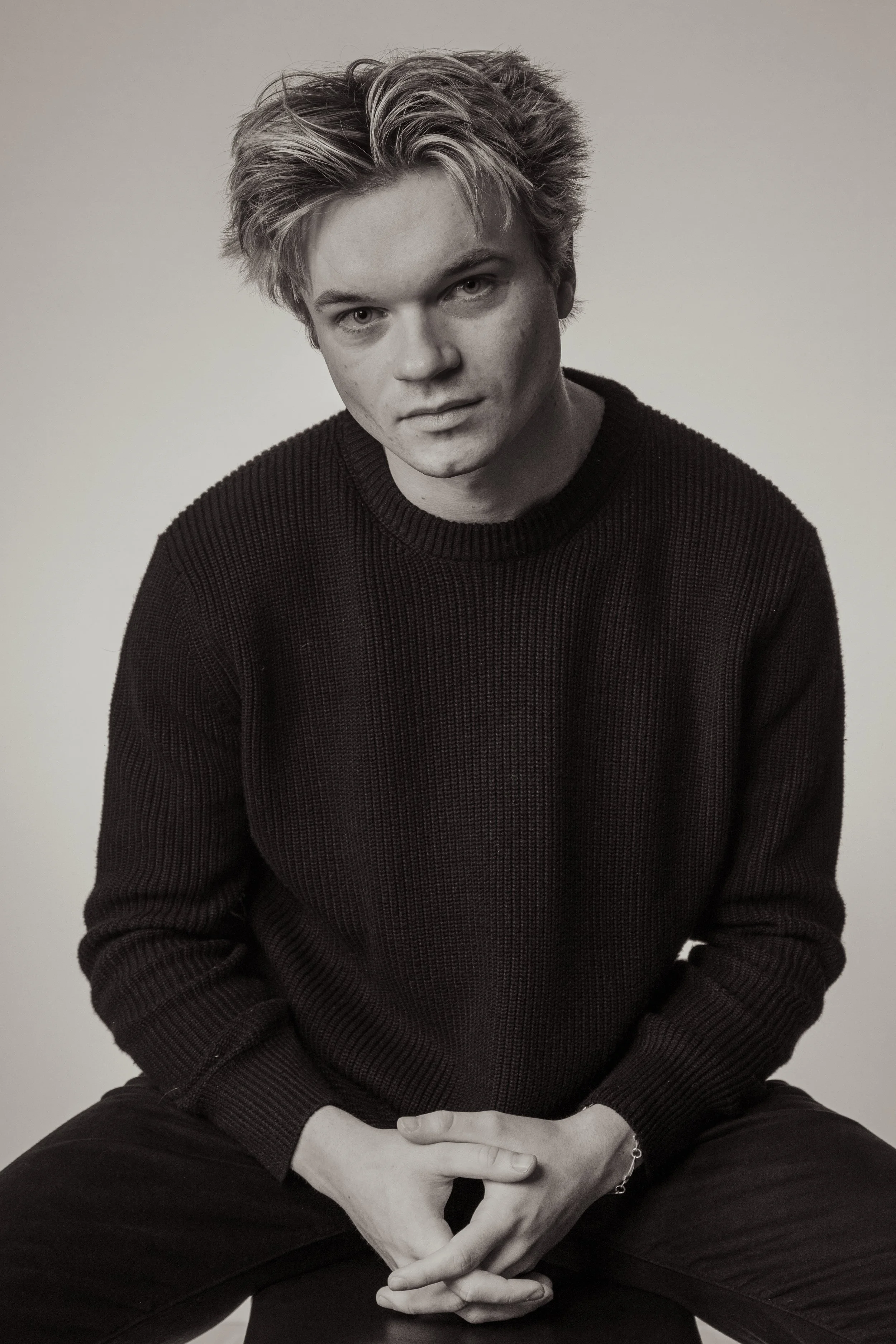 Black and white portrait of a young man with tousled hair, wearing a dark sweater, sitting with hands clasped in front of him against a plain background.