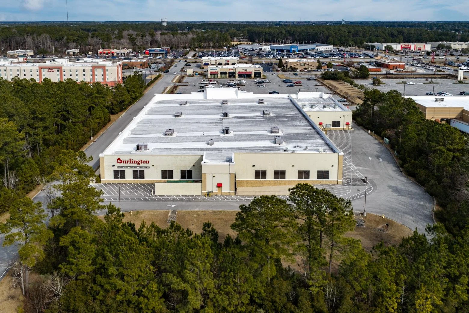 An aerial view of a shopping mall with ample parking lots, surrounded by trees, and a background of retail stores and a forested area under a partly cloudy sky.