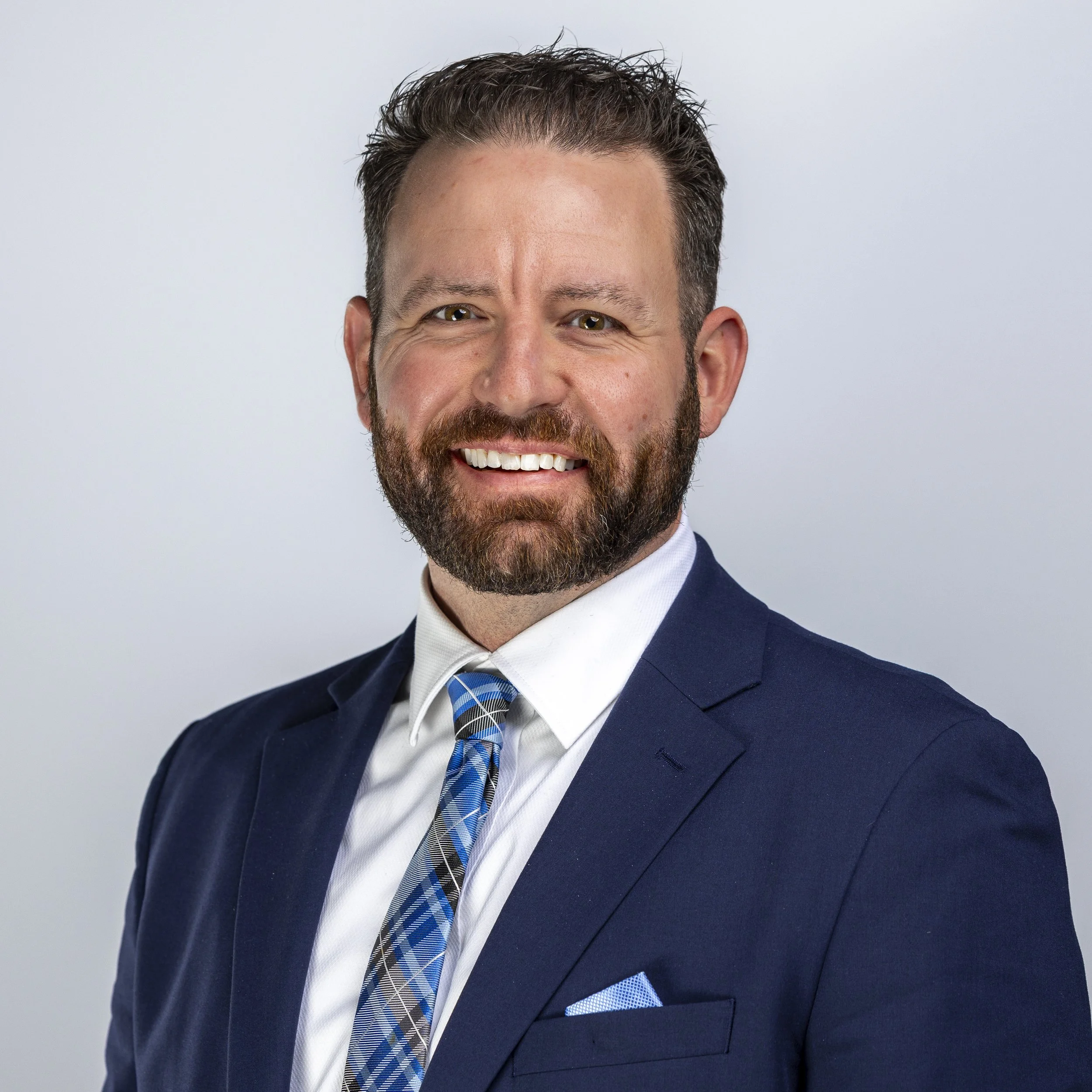 Close-up of a smiling man in a navy suit, white shirt, and blue plaid tie, with a matching blue pocket square, against a plain light gray background.