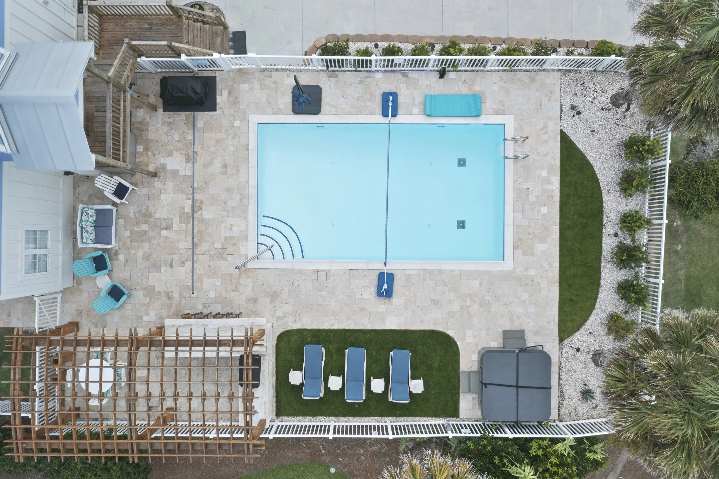Top-down view of a backyard with a swimming pool, pool cleaning equipment, lounge chairs, a small patio area with chairs, ornamental plants, and landscaping.