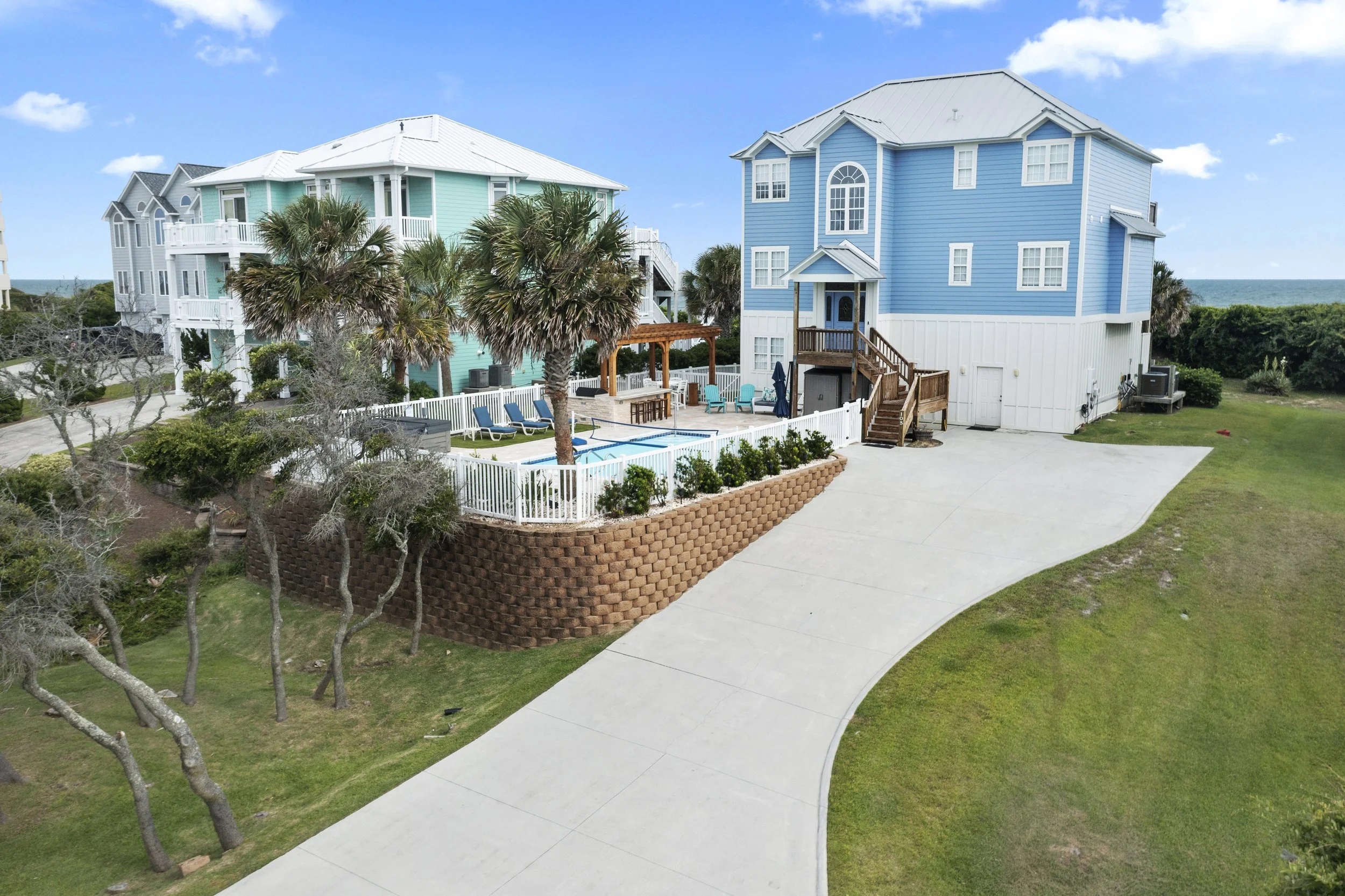Colorful beach house with blue and turquoise exterior, surrounded by palm trees, overlooking the ocean in the background, with a pool area enclosed by a white fence and outdoor furniture.