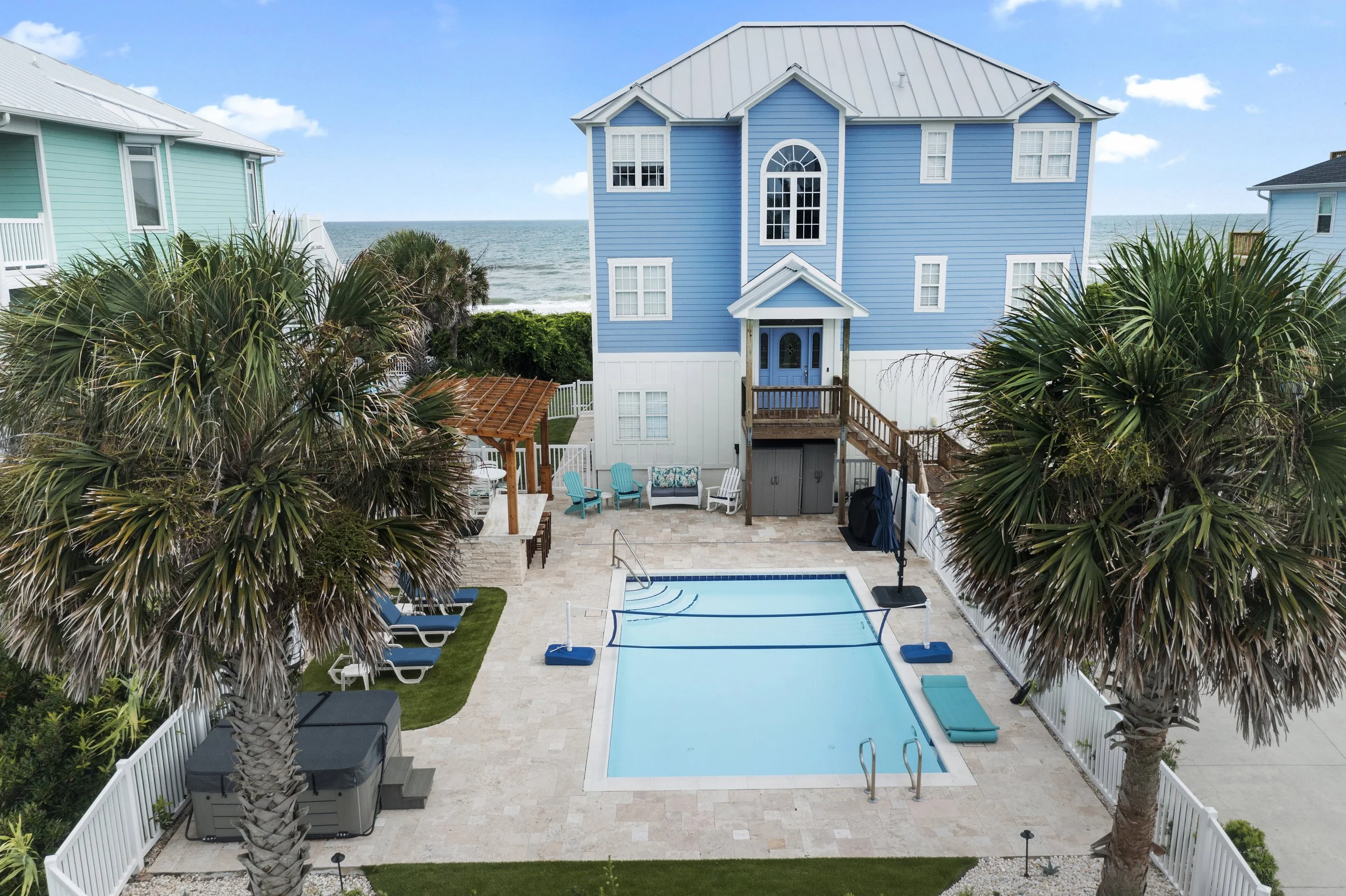 A backyard with a swimming pool, surrounded by palm trees, with a blue house in the background near the beach.