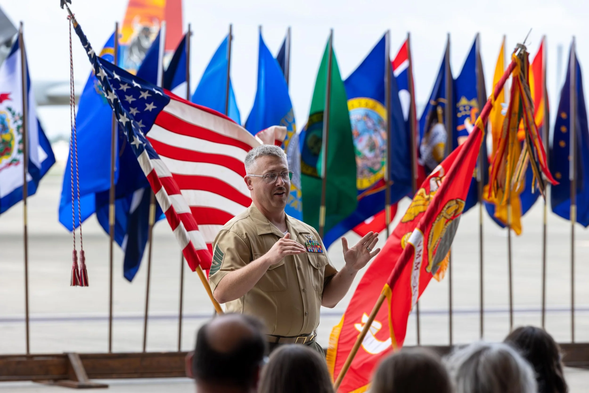 A person in military uniform speaking in front of a display of various American and military flags at an outdoor event.