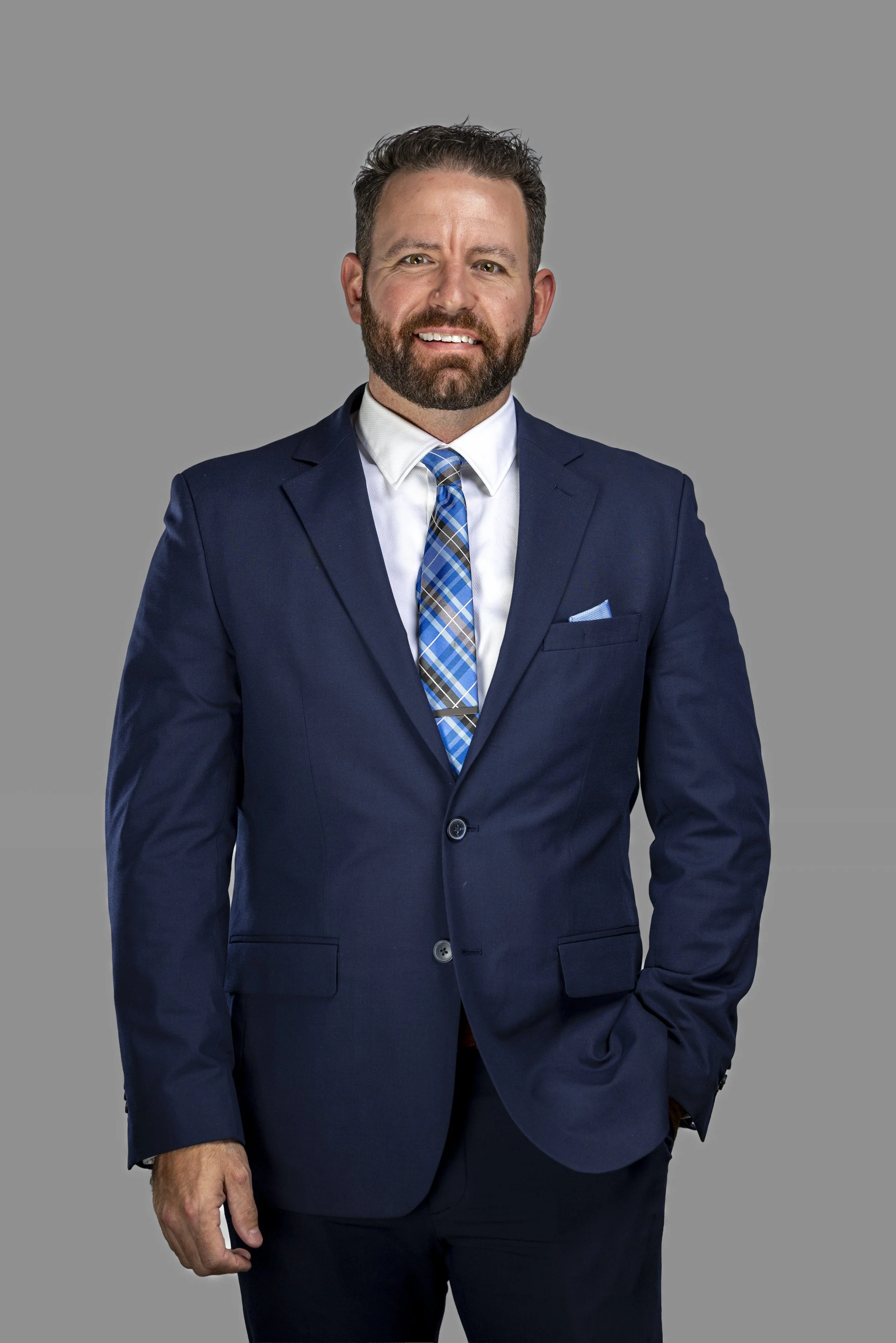 A man with a beard and short hair wearing a navy blue suit, white dress shirt, and a blue plaid tie, standing against a gray background.