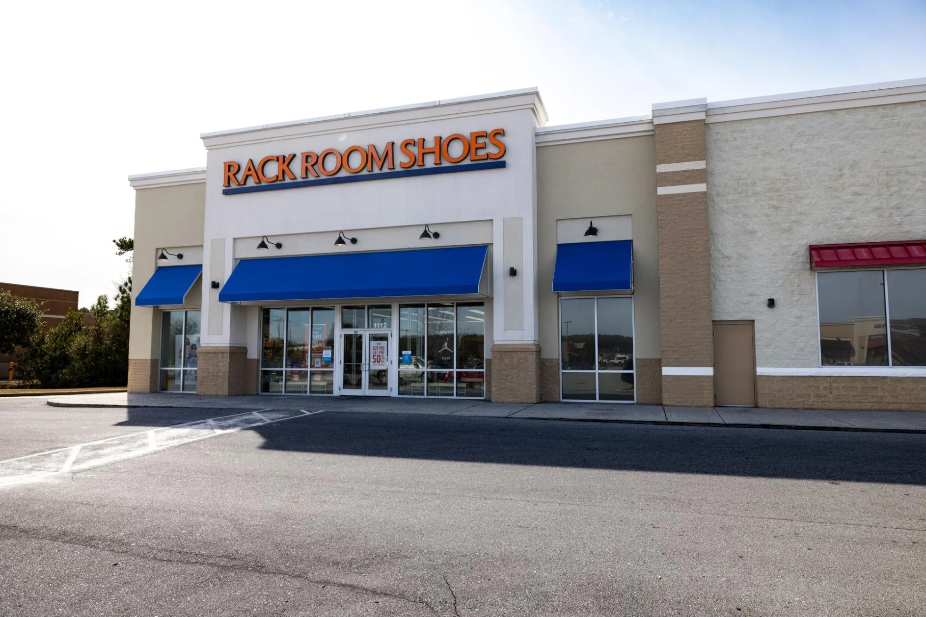Exterior of a retail store called Rack Room Shoes, with blue awnings over the entrance and large glass windows, in a parking lot on a sunny day.