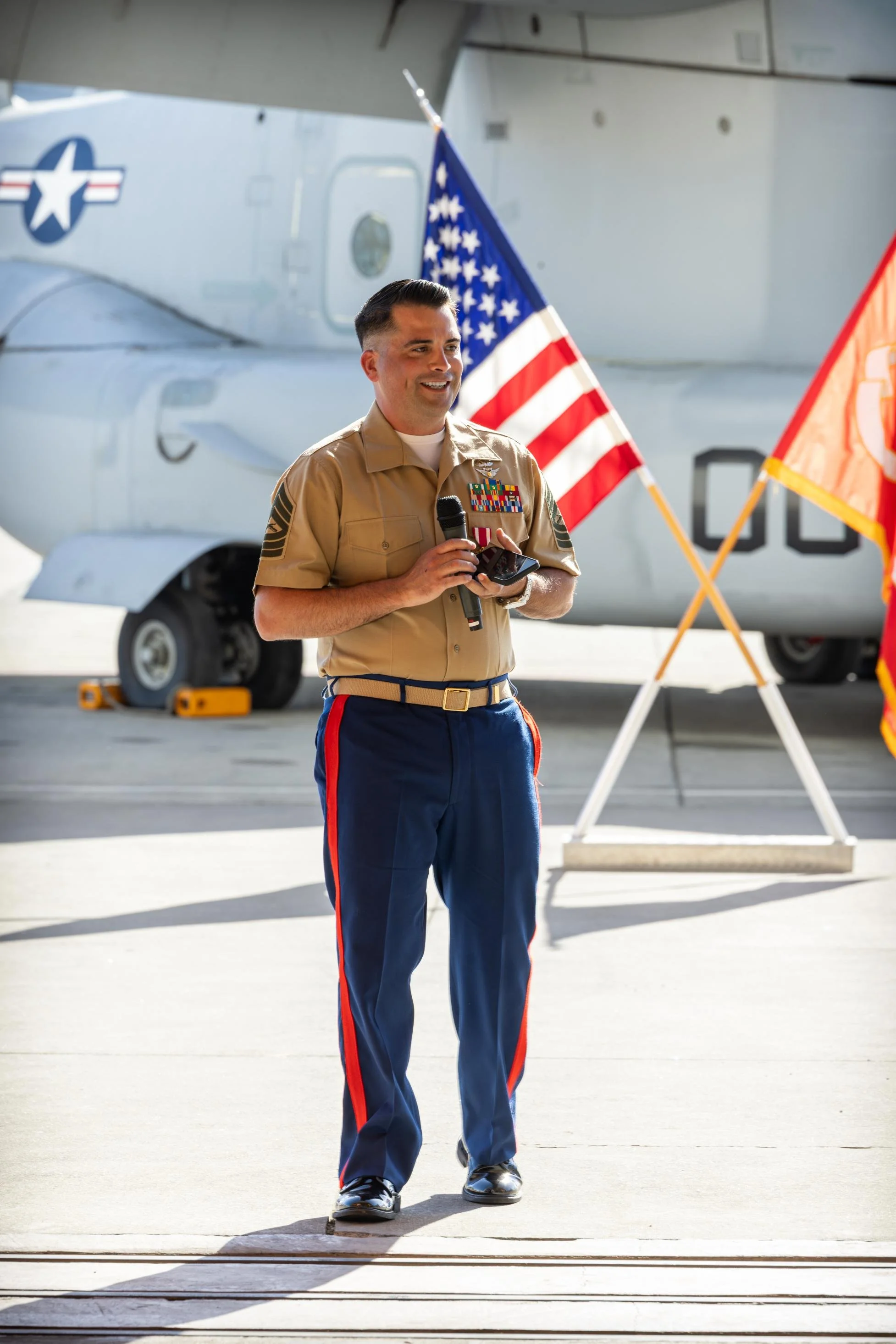 A male military personnel speaking at an event, holding a microphone and a phone, dressed in a tan uniform with medals, standing in front of a military aircraft with American and military flags.