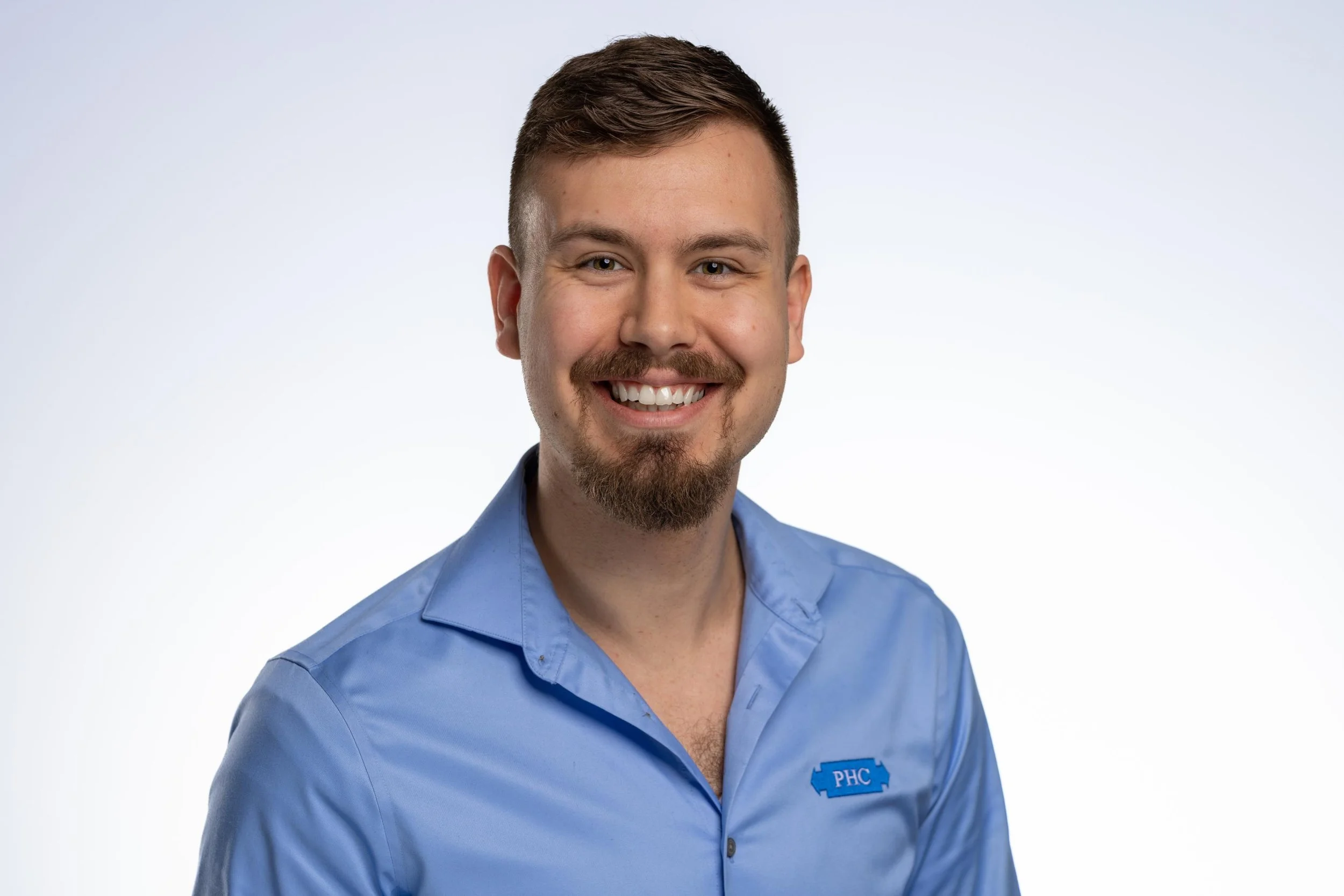 Portrait of a smiling man with short brown hair, a beard, and mustache, wearing a blue collared shirt with a logo on the chest. The background is plain white.