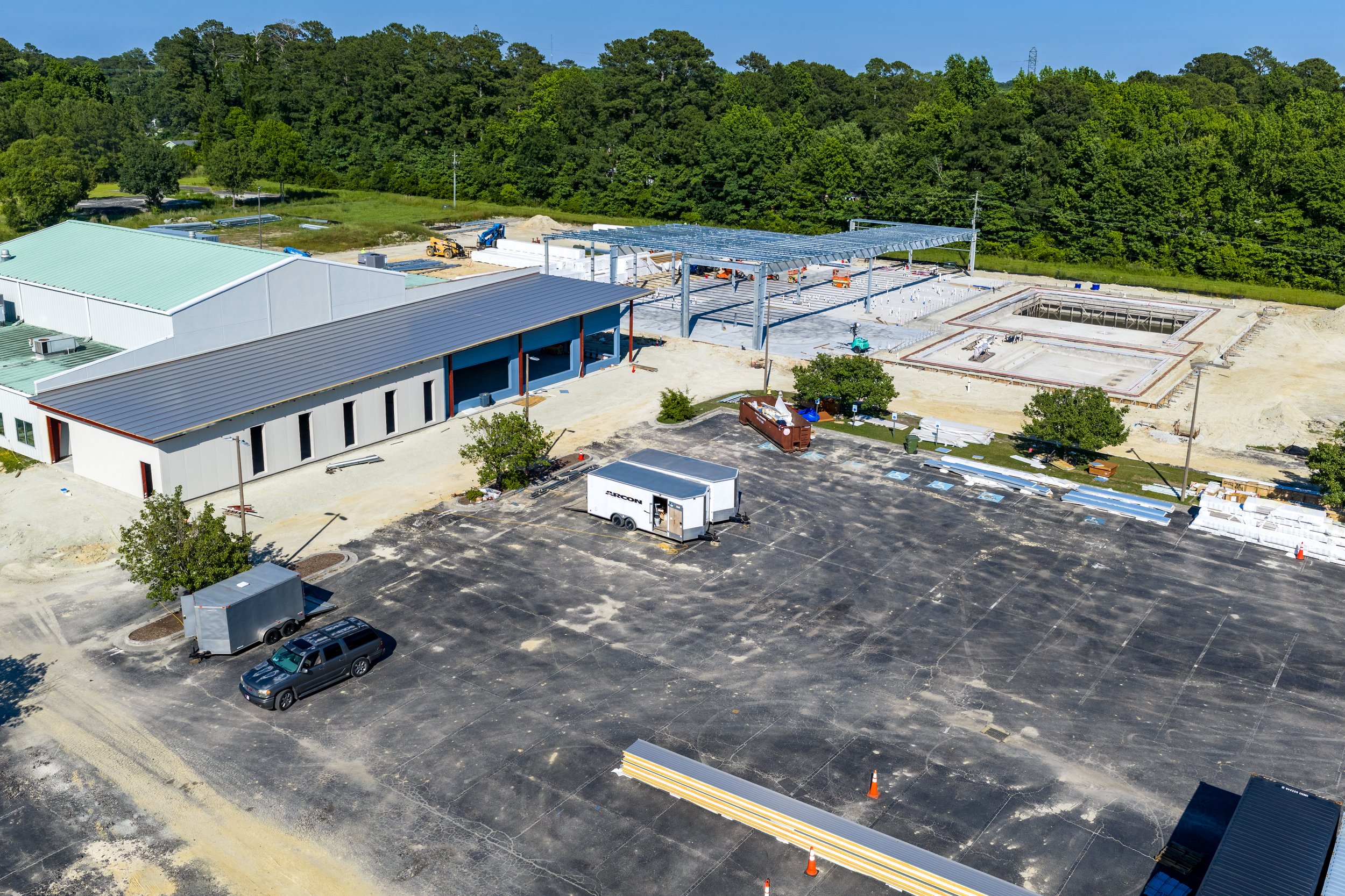 A building under construction with exposed steel framework, construction vehicles, and construction materials surrounding it, with a parking lot and trees in the background.