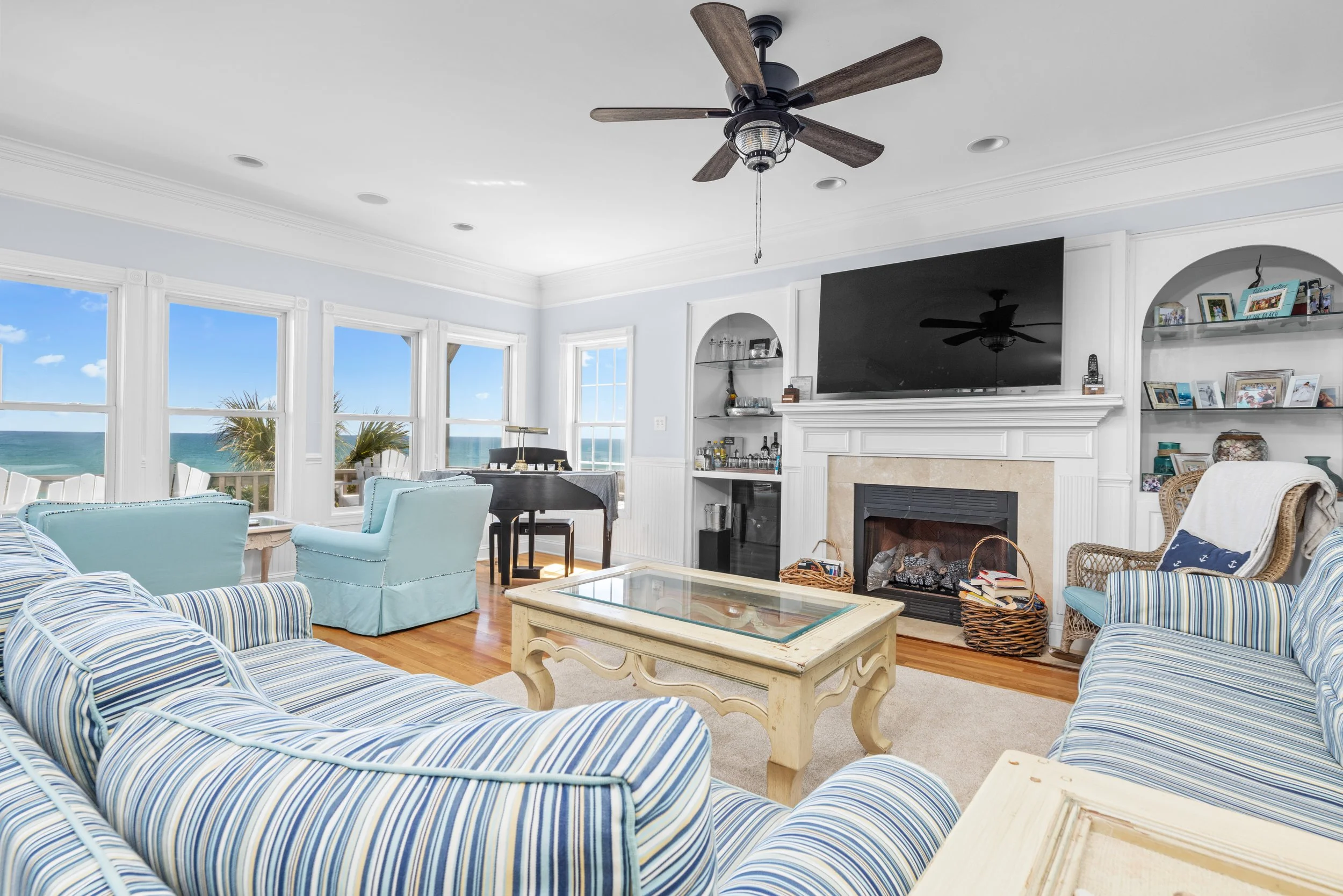 Living room with large windows showing ocean view, light blue and striped sofas, wooden coffee table, ceiling fan, fireplace, shelves with photos and decor.