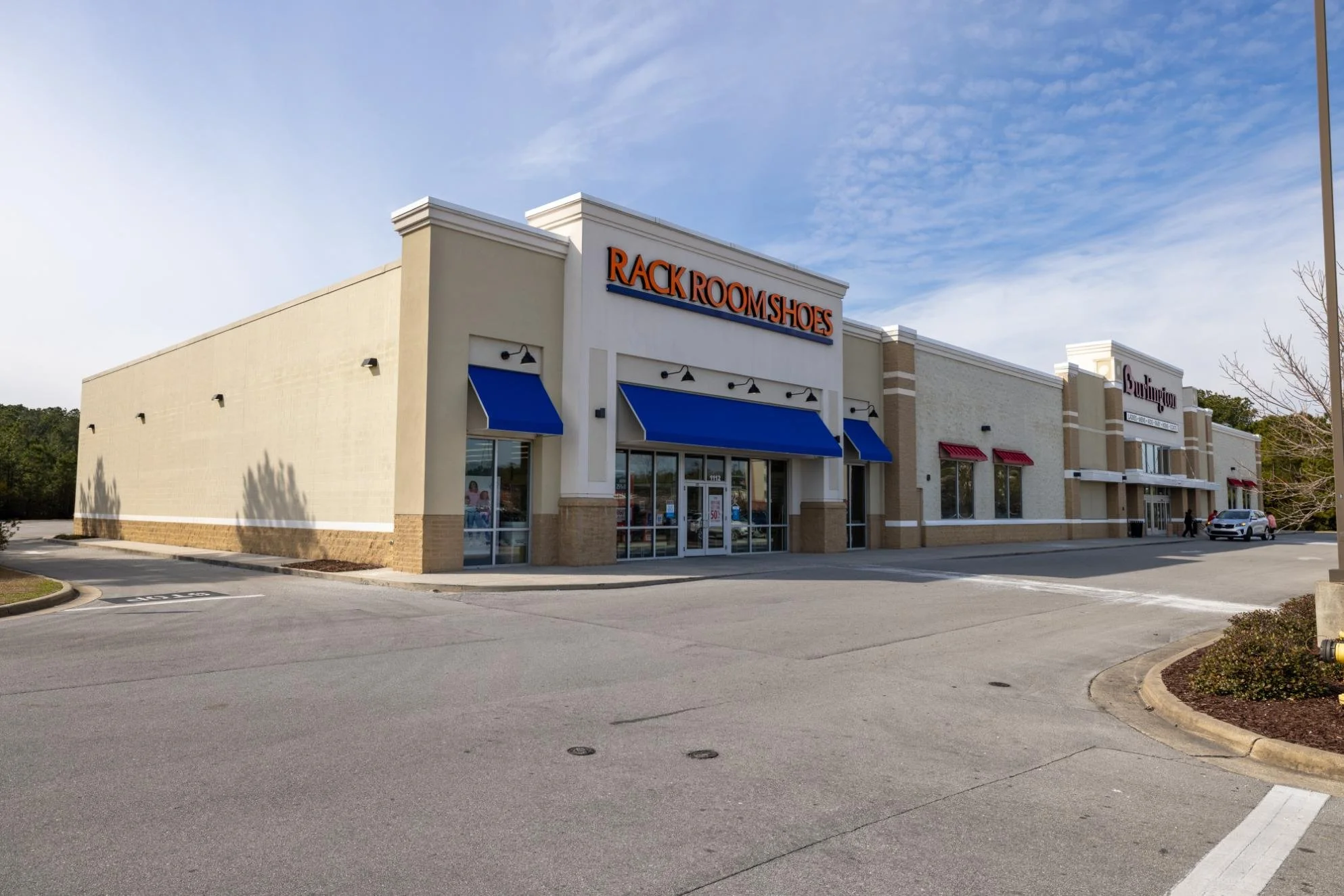 Shopping plaza with a store called Rack Room Shoes and another store, Burlington, with both stores having blue and red awnings respectively. The parking lot is mostly empty with a few cars and pedestrians walking nearby, and the sky is partly cloudy.