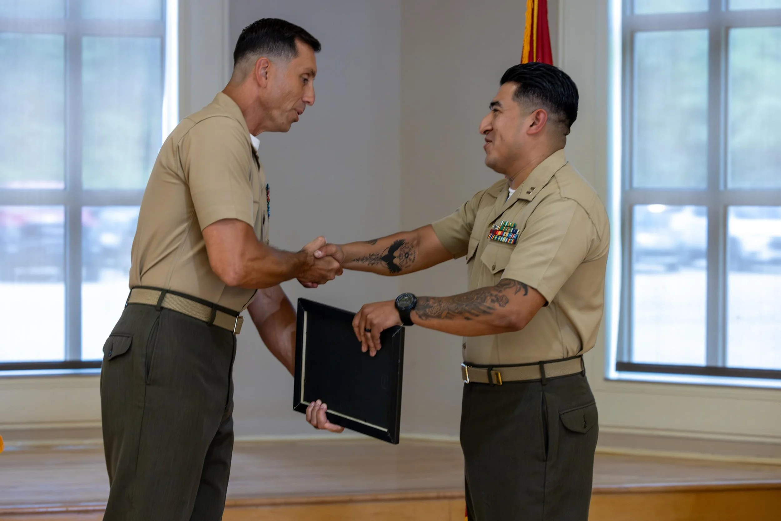 Two military officers shaking hands, one presenting an award or plaque indoors with large windows and a flag in the background.