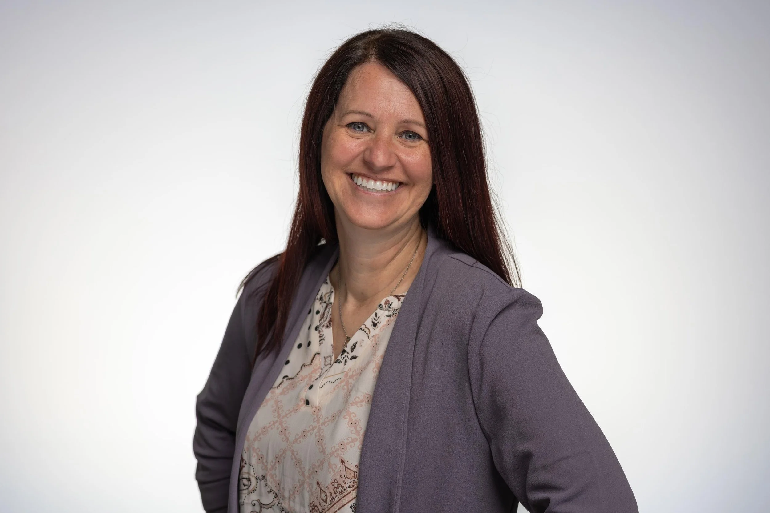 A woman with long dark hair and light skin smiling at the camera, wearing a gray blazer over a patterned blouse, set against a plain white background.