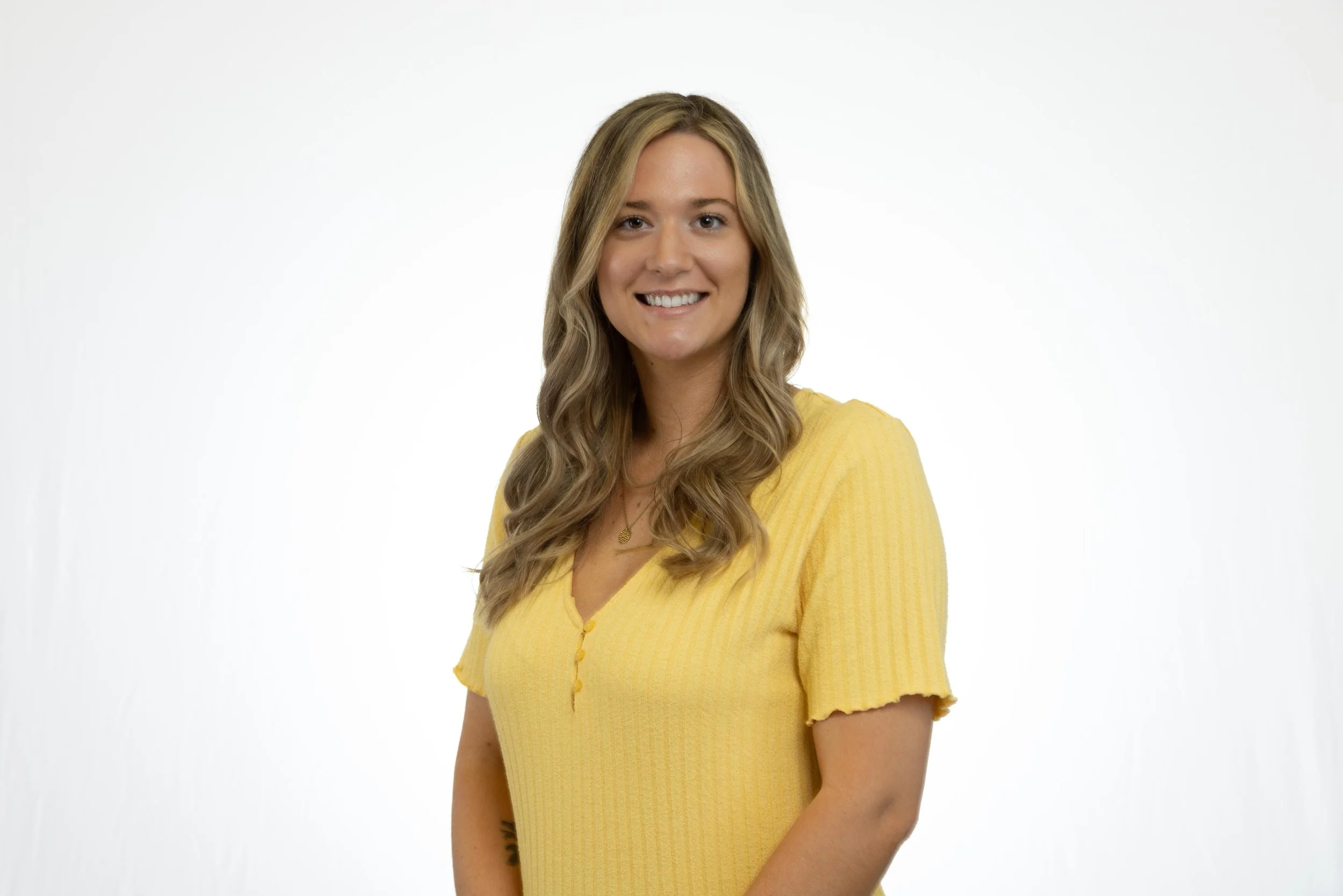 A woman with long wavy blonde hair smiling, wearing a yellow short-sleeved, button-up top, standing against a plain white background.