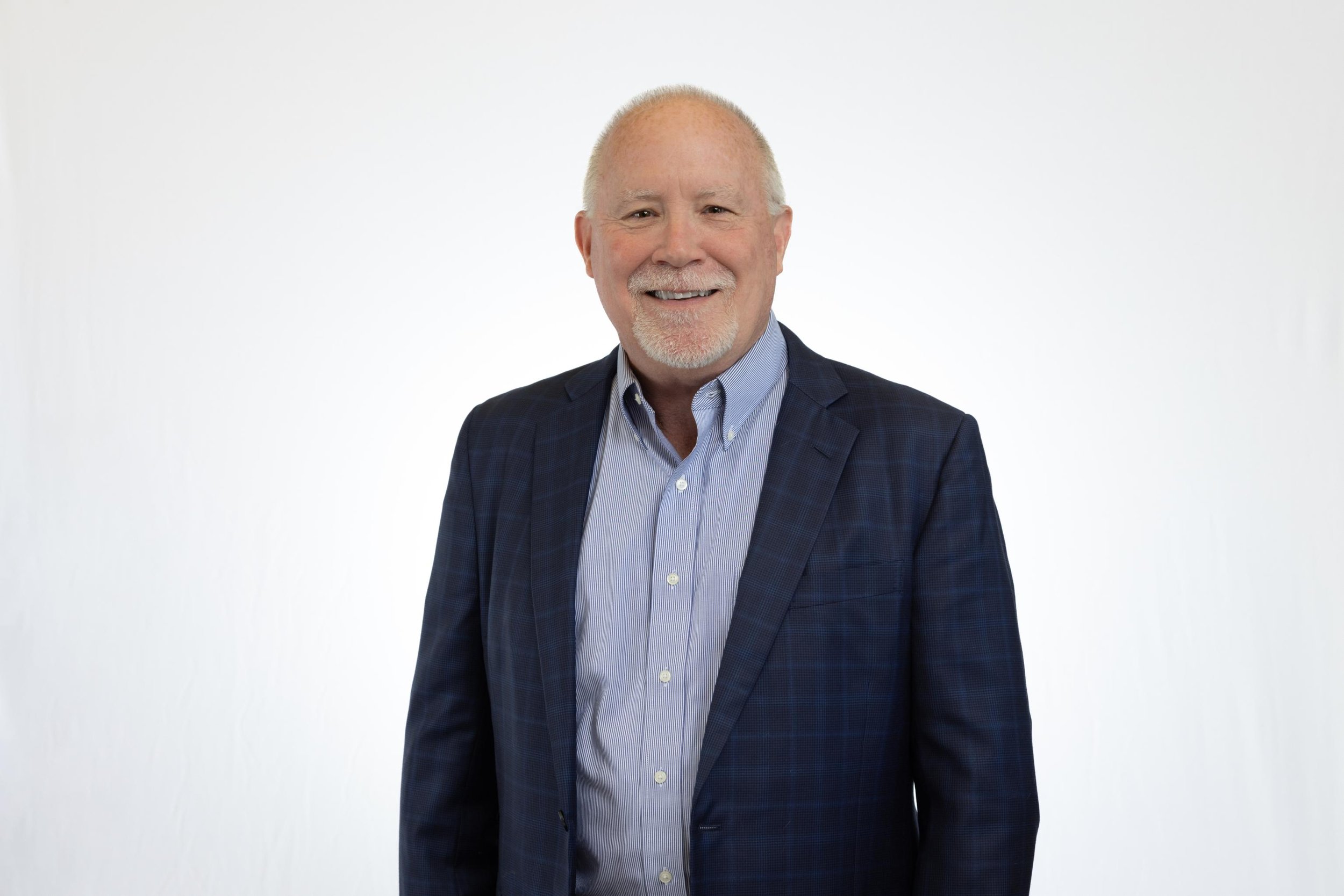 Portrait of a smiling older man with short gray hair and a beard, wearing a dark plaid blazer and a light blue dress shirt, standing against a plain white background.