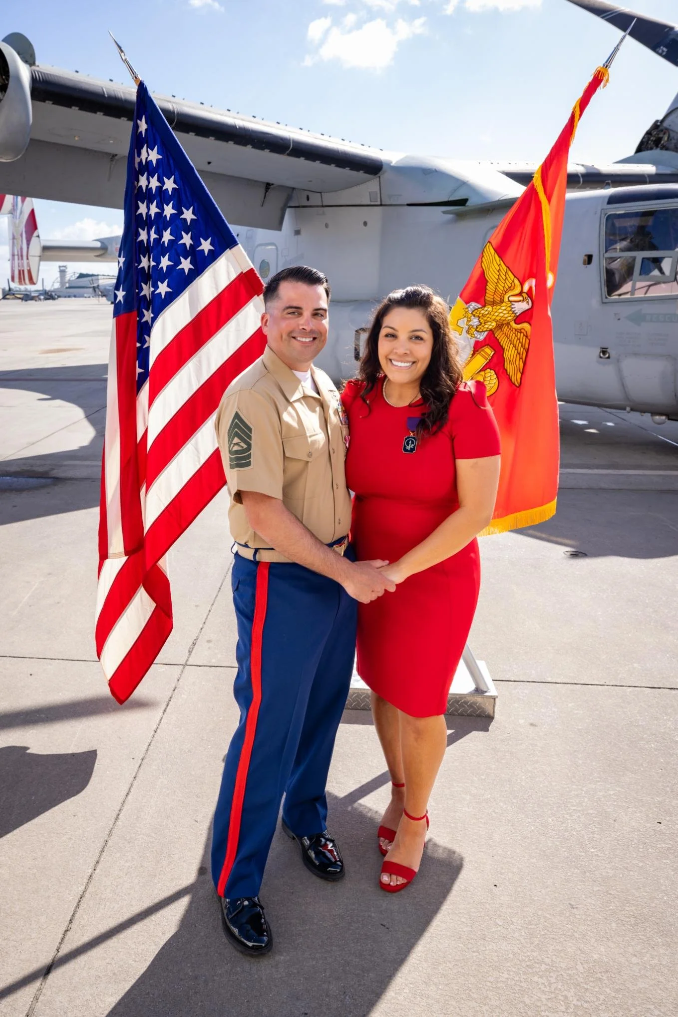 A man in a military uniform and a woman in a red dress holding hands, standing in front of a military aircraft with American and Marine flags.