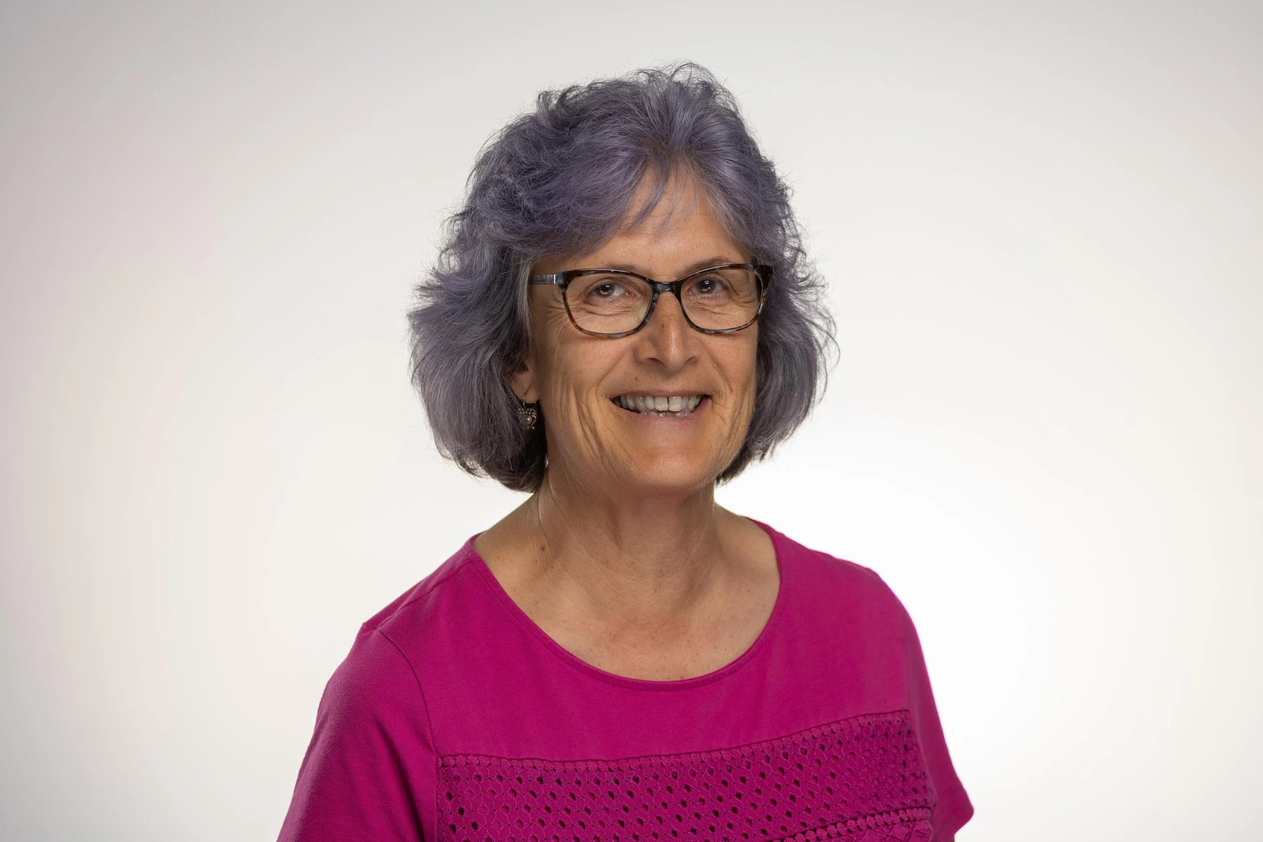 A middle-aged woman with short gray hair and glasses smiling at the camera, wearing a pink shirt with a textured pattern on the front, standing against a plain white background.
