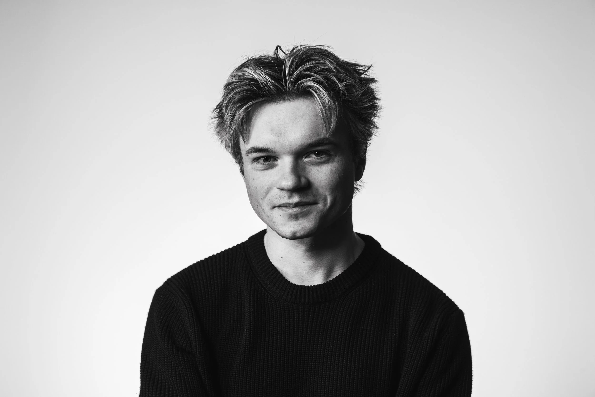 Black and white portrait of a young man with tousled hair, wearing a dark sweater, looking directly at the camera, against a plain light background.