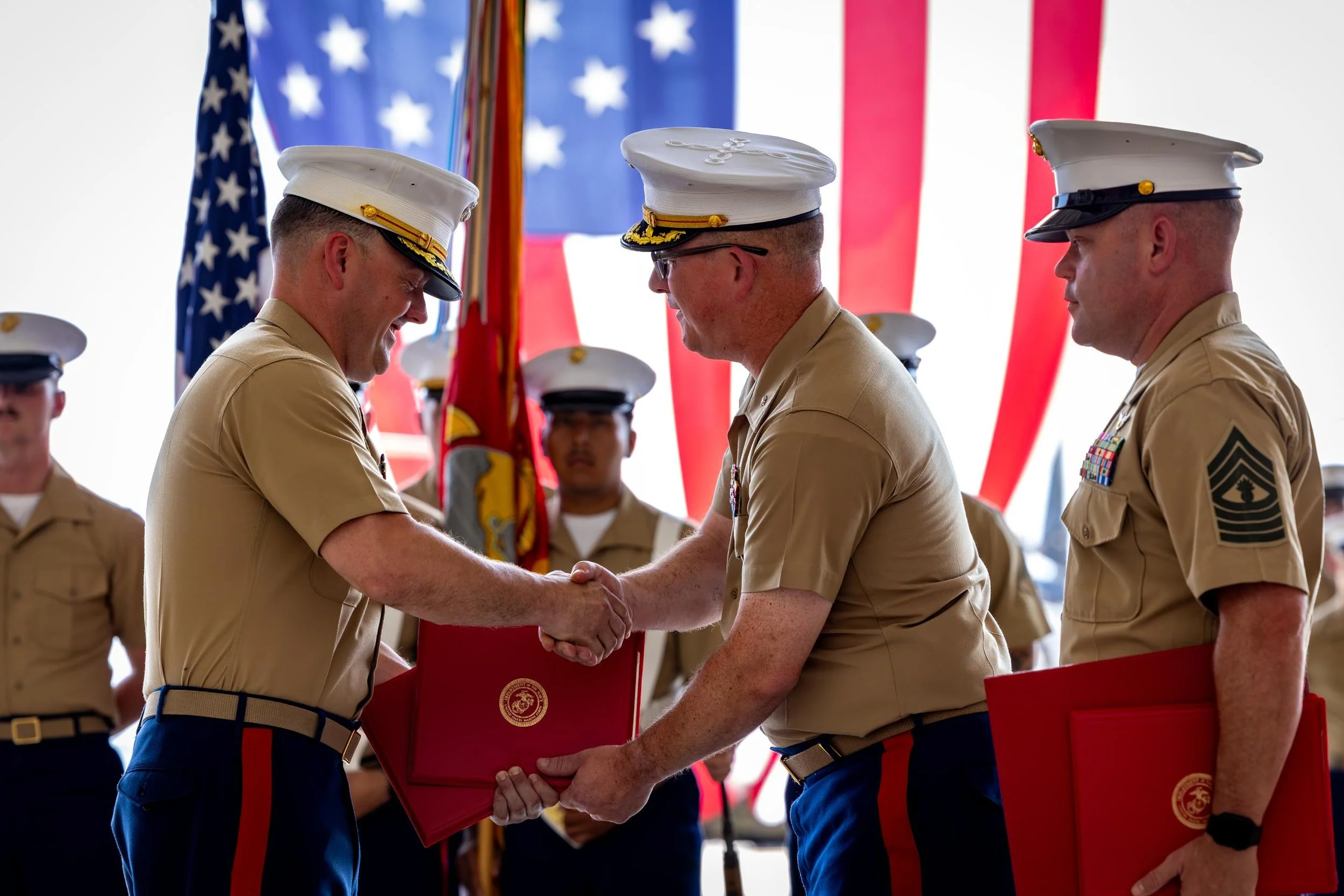 Military officers in uniform shaking hands during a formal ceremony with American flags in the background.