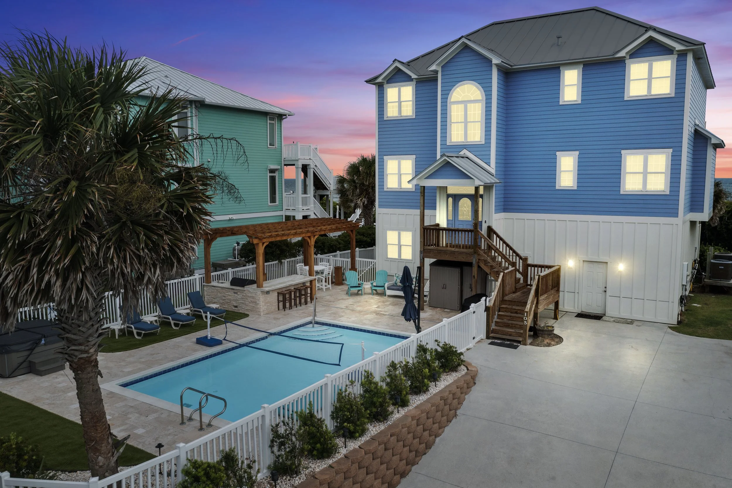 A backyard scene with two colorful houses, one painted blue and the other green, overlooking a swimming pool area with lounge chairs, a bar, and outdoor furniture, surrounded by a white fence and palm trees during sunset.