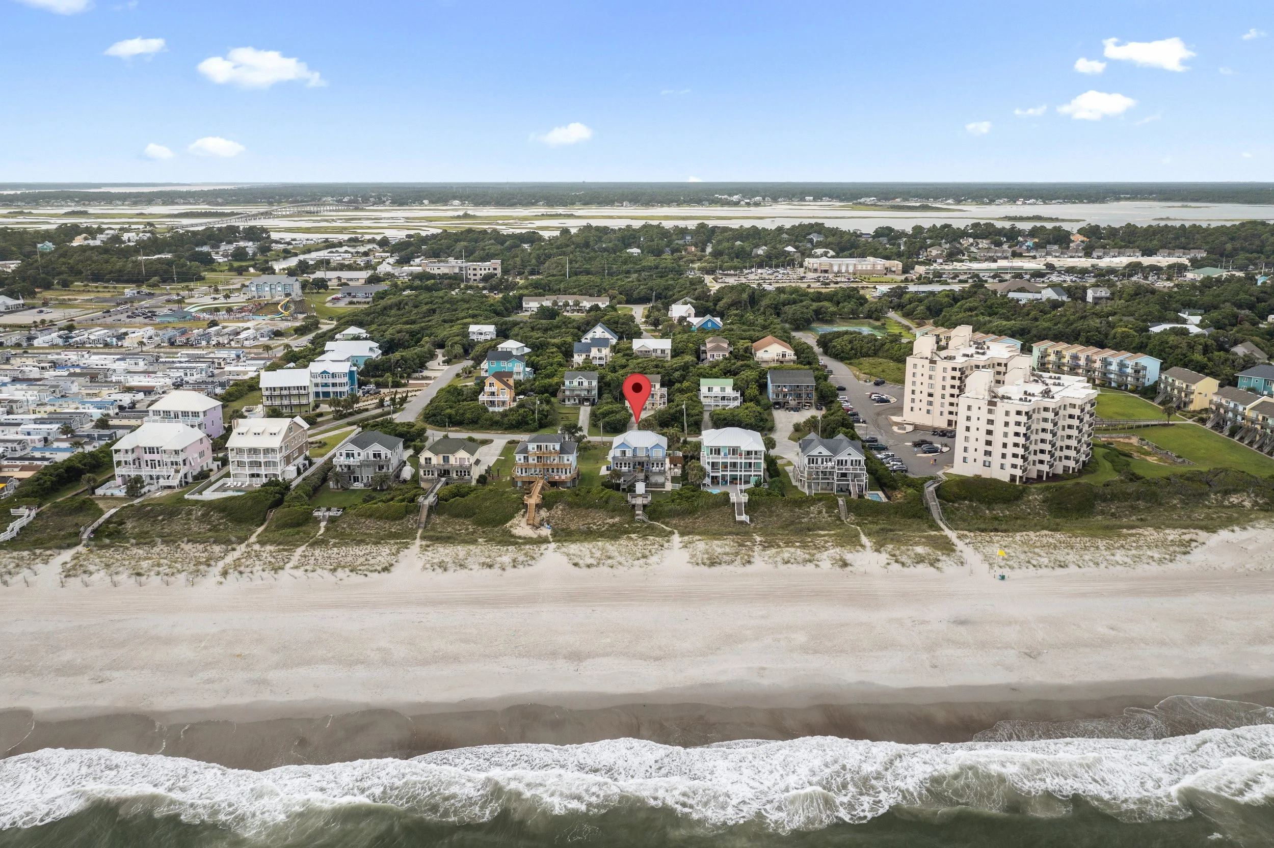 Aerial view of a beachside neighborhood with colorful houses, green trees, and a sandy shoreline, overlooking the ocean and distant waters under a blue sky with scattered clouds.