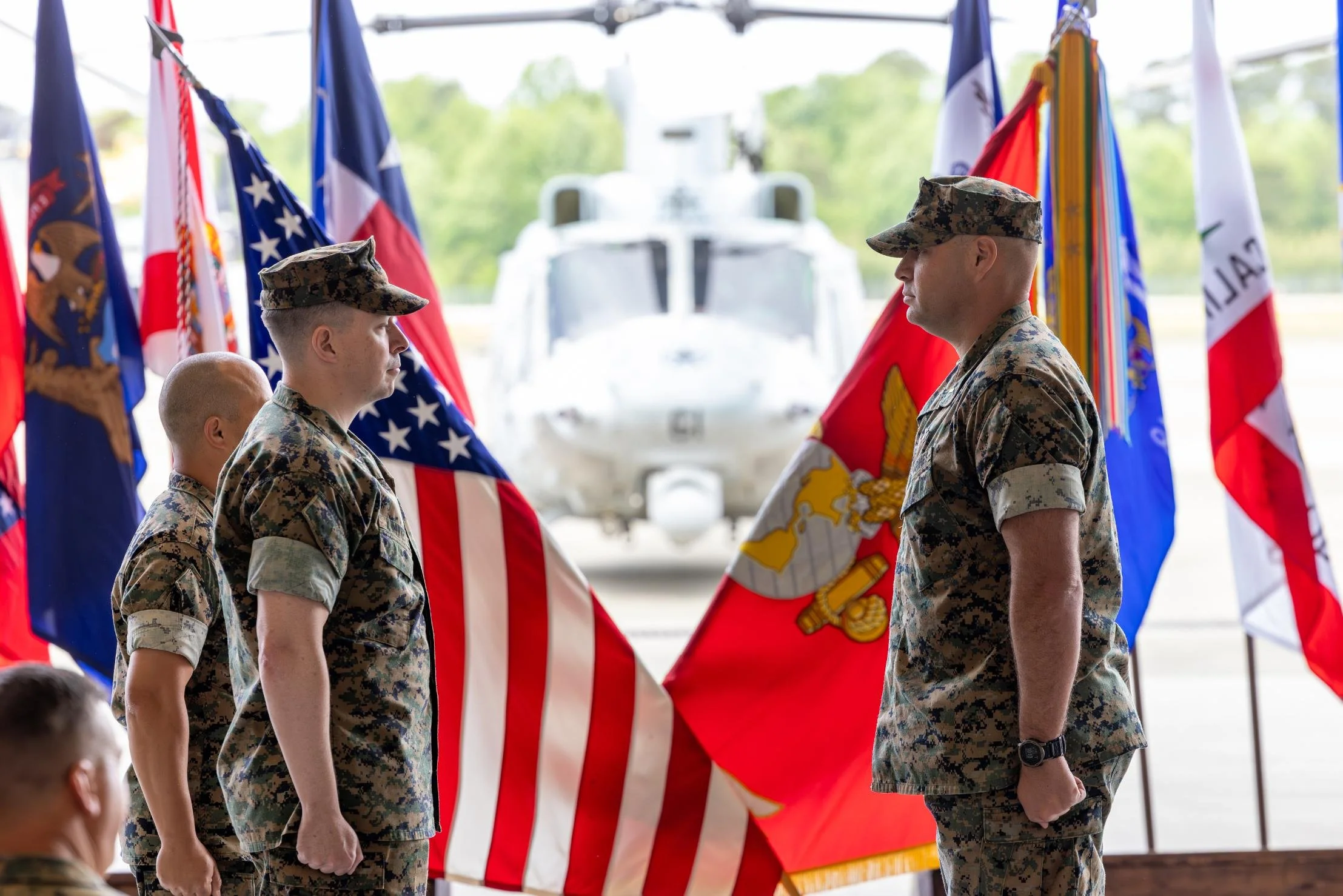 Two soldiers in camouflage uniforms and hats stand face-to-face during a military ceremony, with a large helicopter and multiple flags in the background.