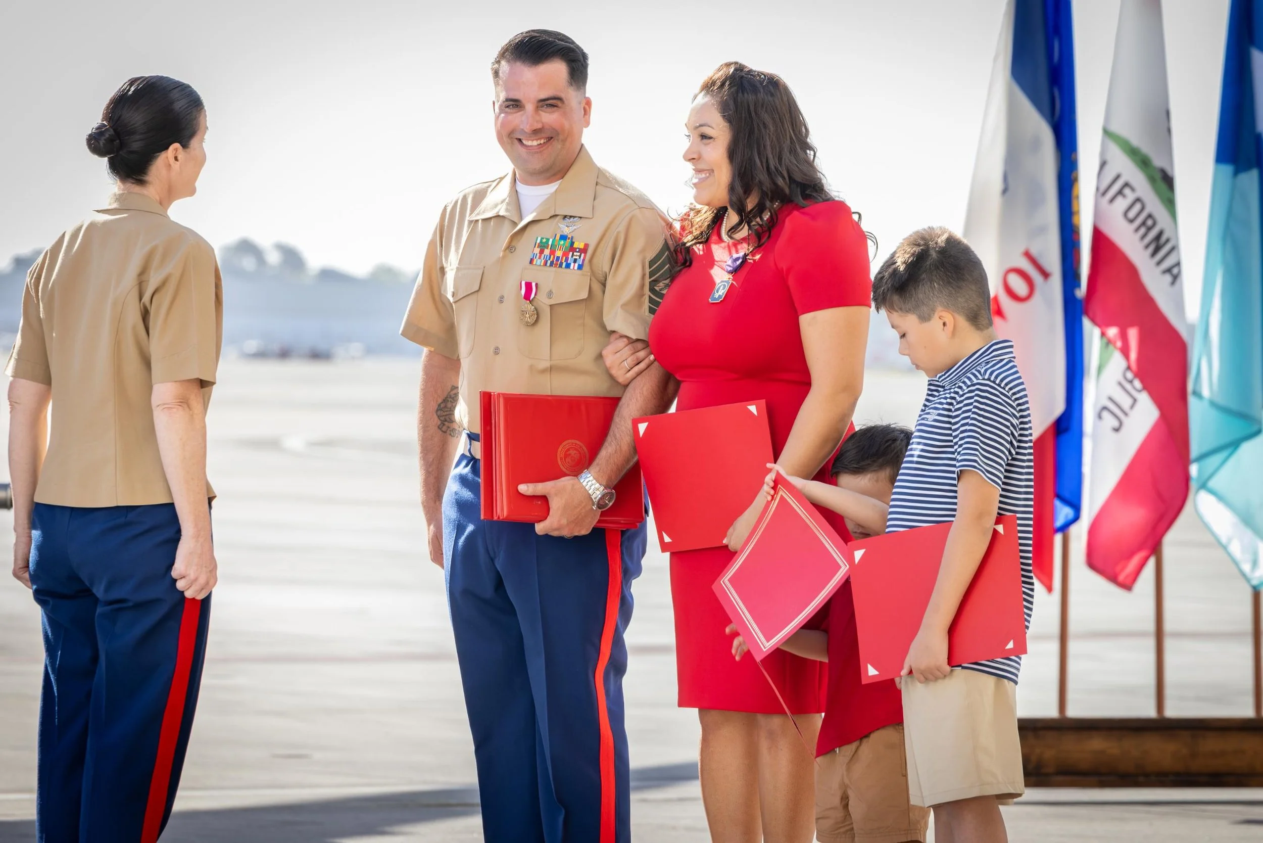 A family at an airport outside gate, holding red certificates, looking proud as military officers in uniform shake hands with them. Flags are visible in the background.