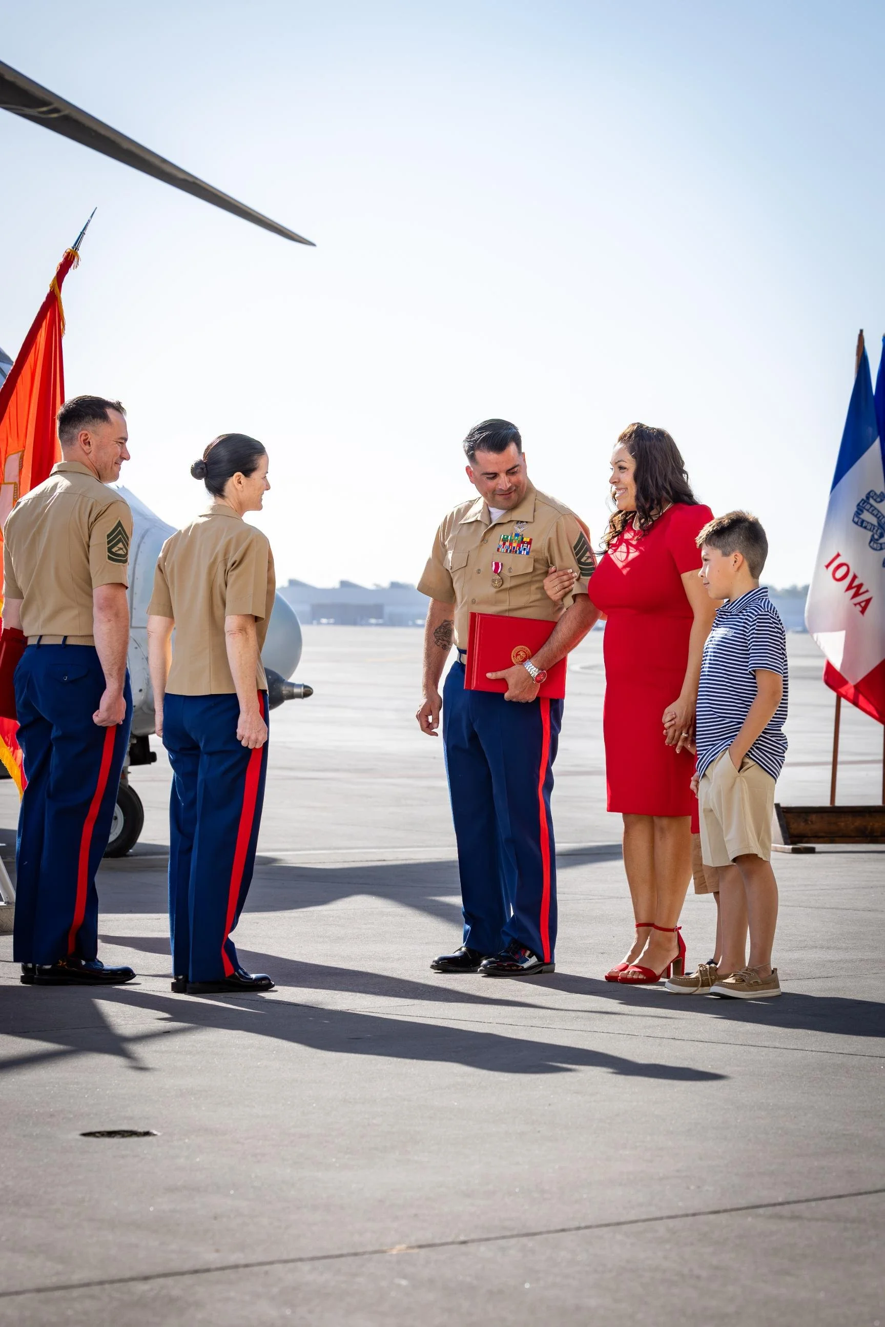 A group of people, including military personnel in uniform, a woman in a red dress, and a young boy, standing on an airport tarmac with flags in the background. The scene appears to be a welcoming or farewell ceremony.