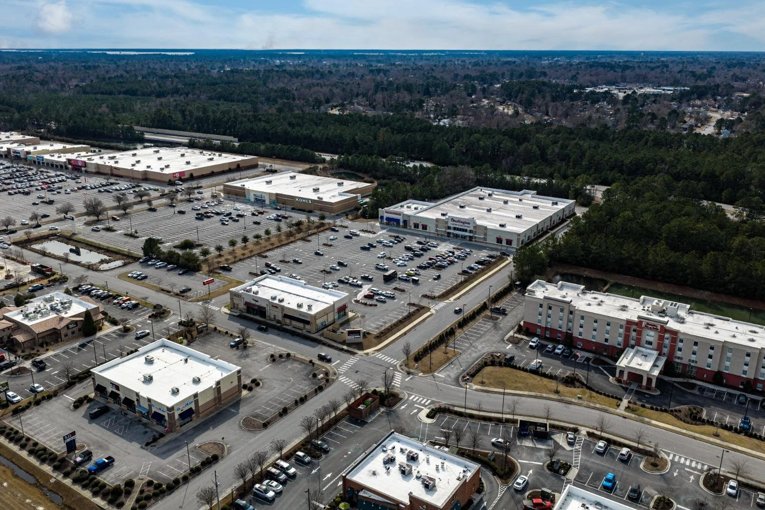 An aerial view of a shopping center with large parking lots, surrounded by trees and distant landscape.