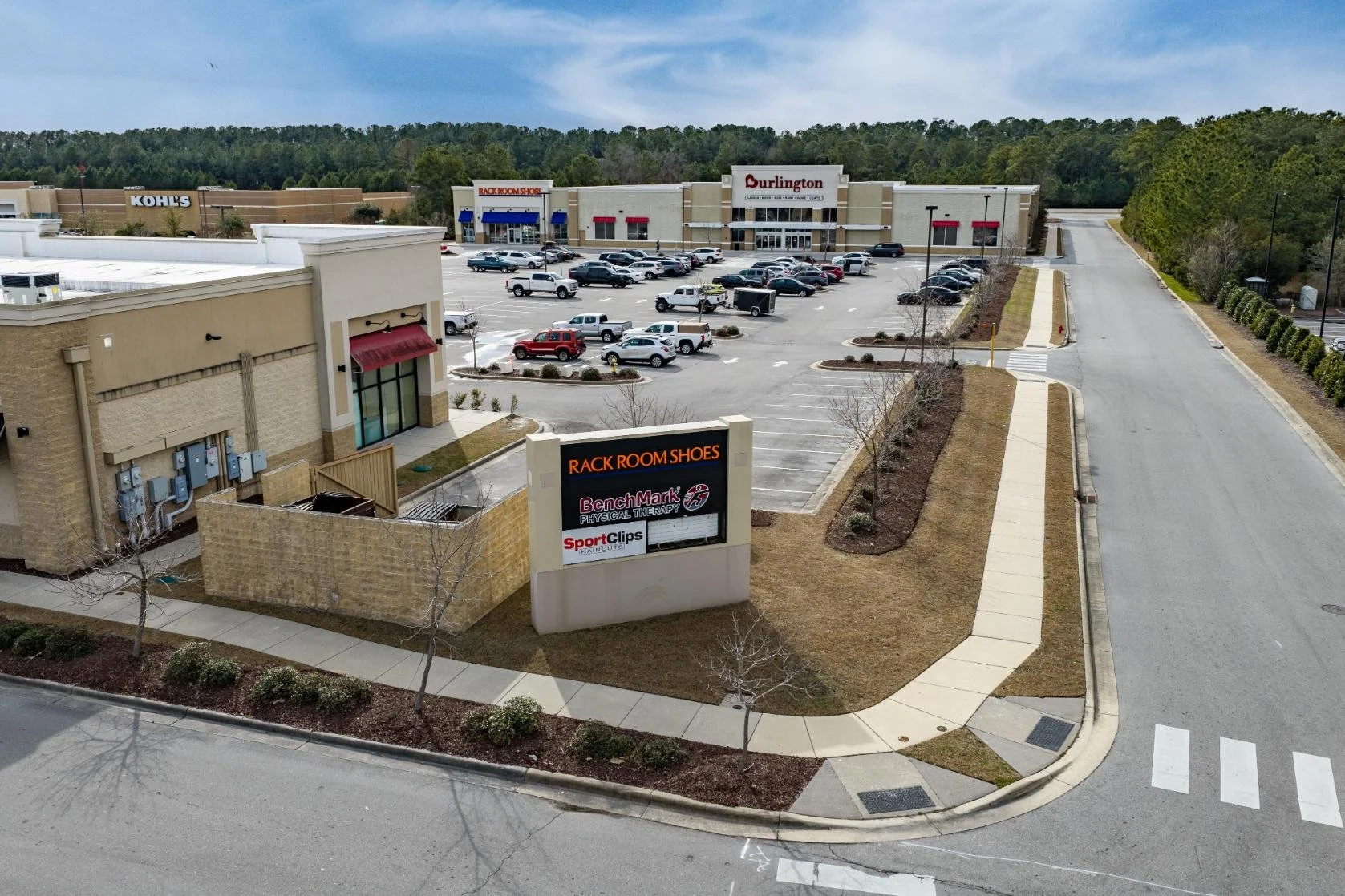 A shopping plaza with stores including Rack Room Shoes, Burlington, Kohl's, and other shops, with a parking lot full of cars and a road with a sidewalk in a suburban area. There are trees and a wooded hill in the background.
