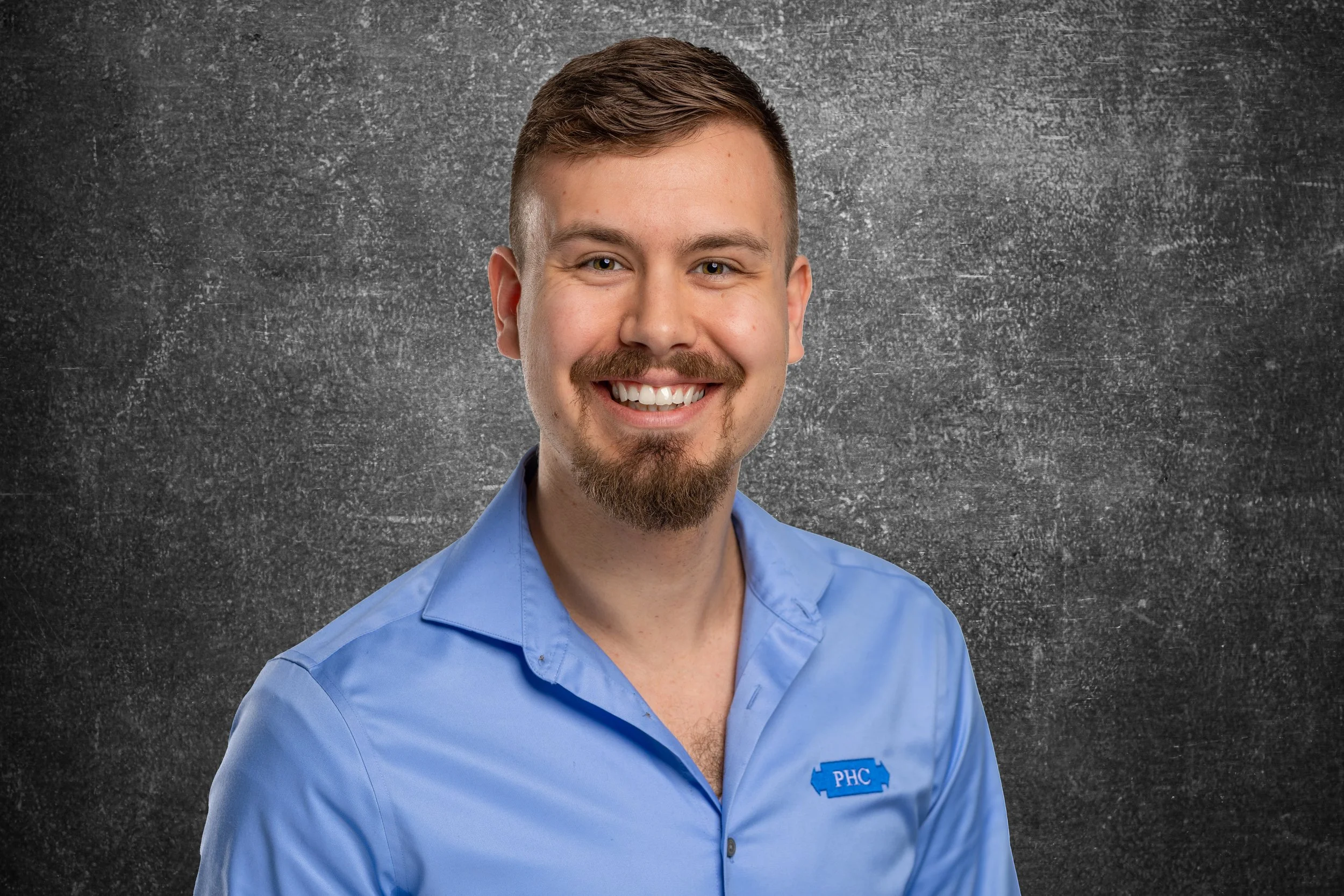 A young man with short brown hair, a goatee, and mustache, smiling, wearing a blue button-down shirt with an embroidered patch that says "PHC". He is standing against a dark gray textured background.