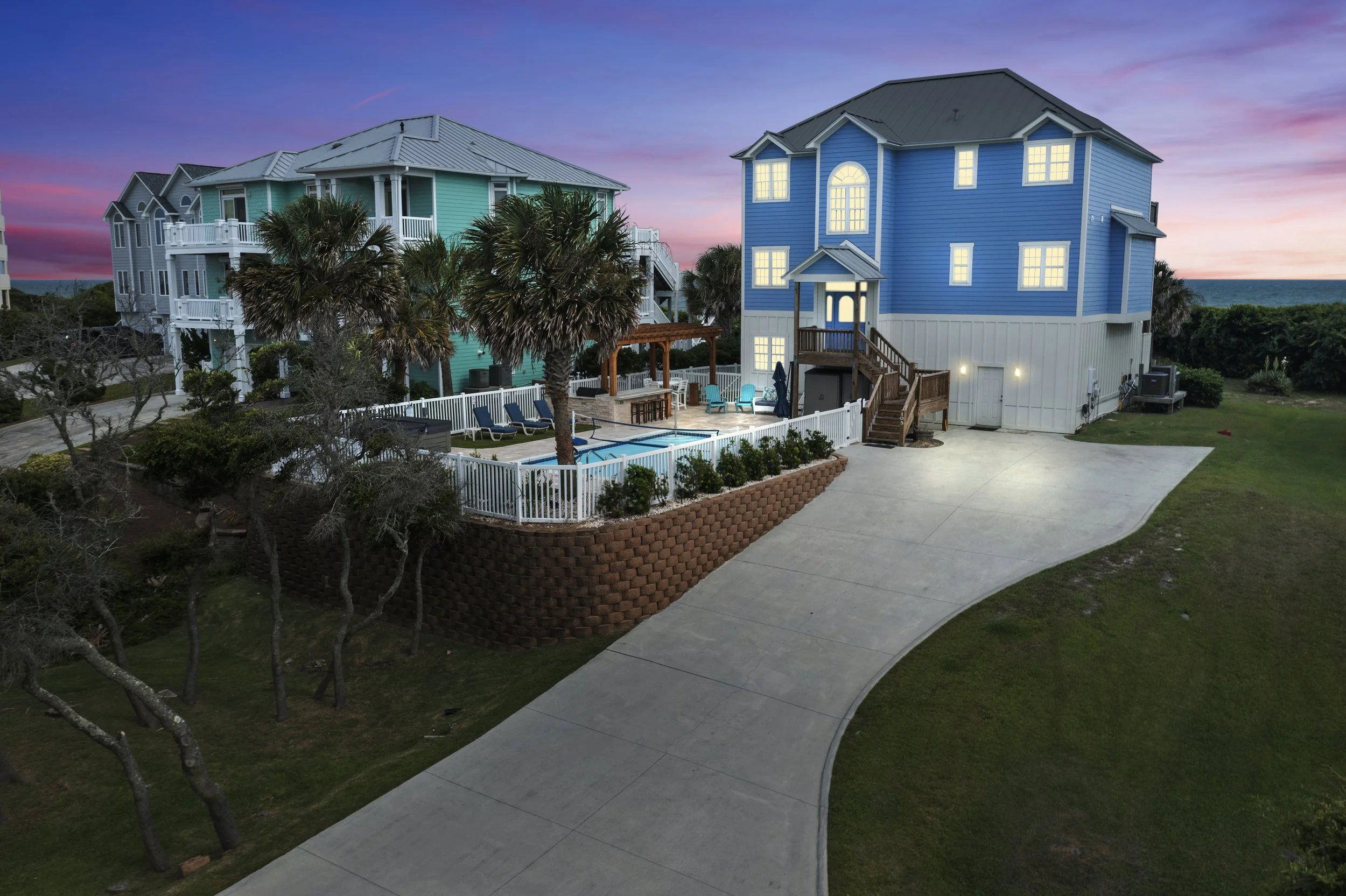 A seaside residential area during sunset featuring multi-story blue and teal houses with white railings, palm trees, a swimming pool, and a driveway, with the ocean visible in the background.