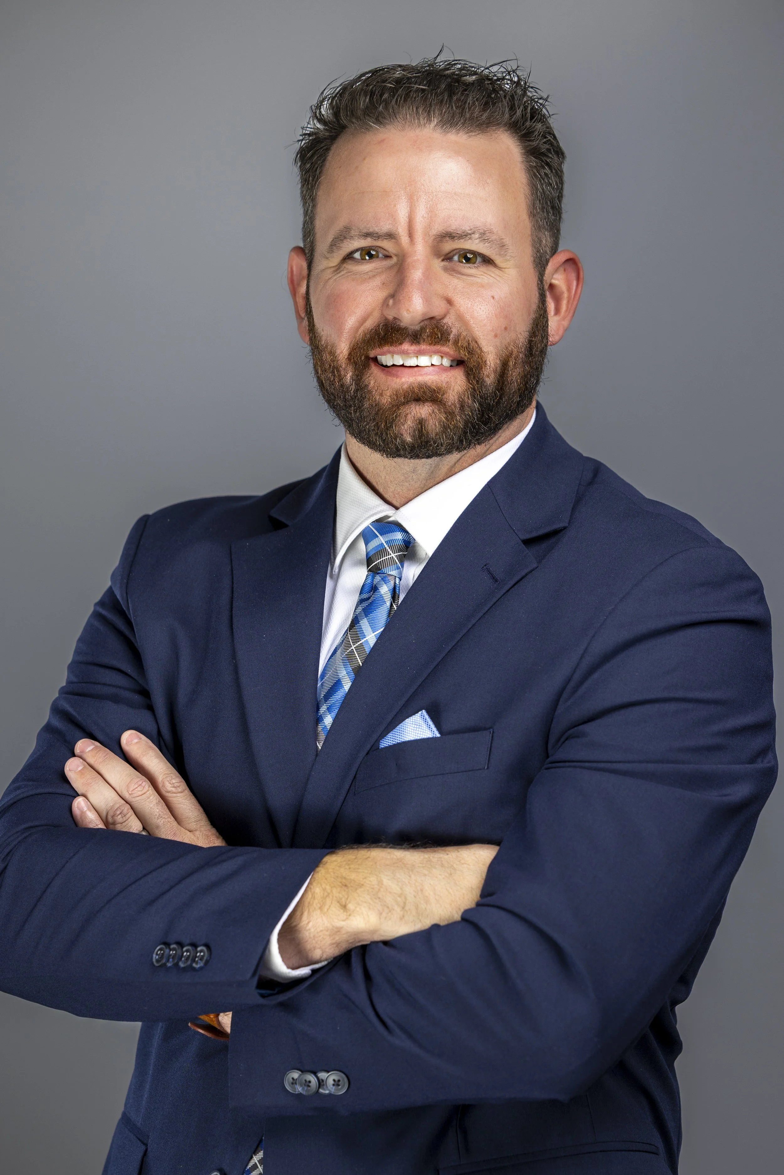A smiling man with a beard and mustache wearing a navy blue suit, white shirt, and blue patterned tie, with arms crossed, standing against a gray background.