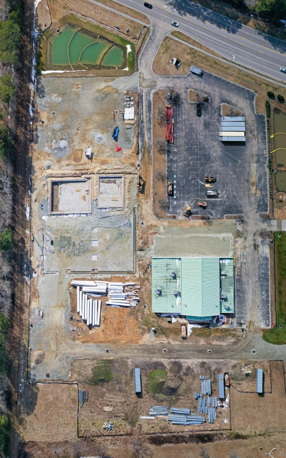 Aerial view of a construction site with foundation work, construction materials, and vehicles, bordered by a parking lot and a road on the right side.