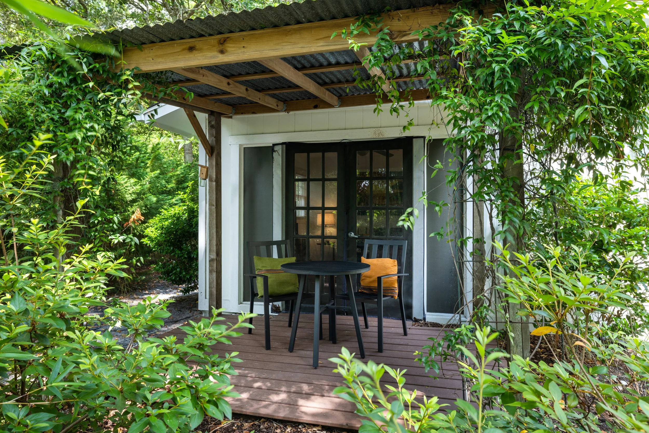 A small outdoor patio with black chairs and orange and green cushions, surrounded by green leafy plants. There is a white wall with a black window and a wooden deck floor. The patio has a roof made of corrugated metal and wooden beams.