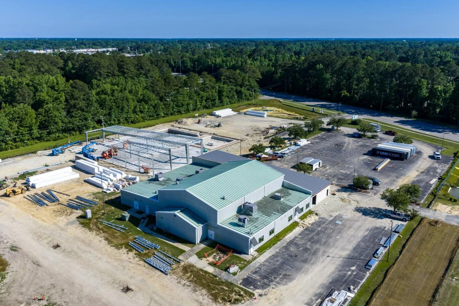 Construction site with a large building under construction, construction vehicles, and materials, surrounded by a parking lot and wooded area in the background.