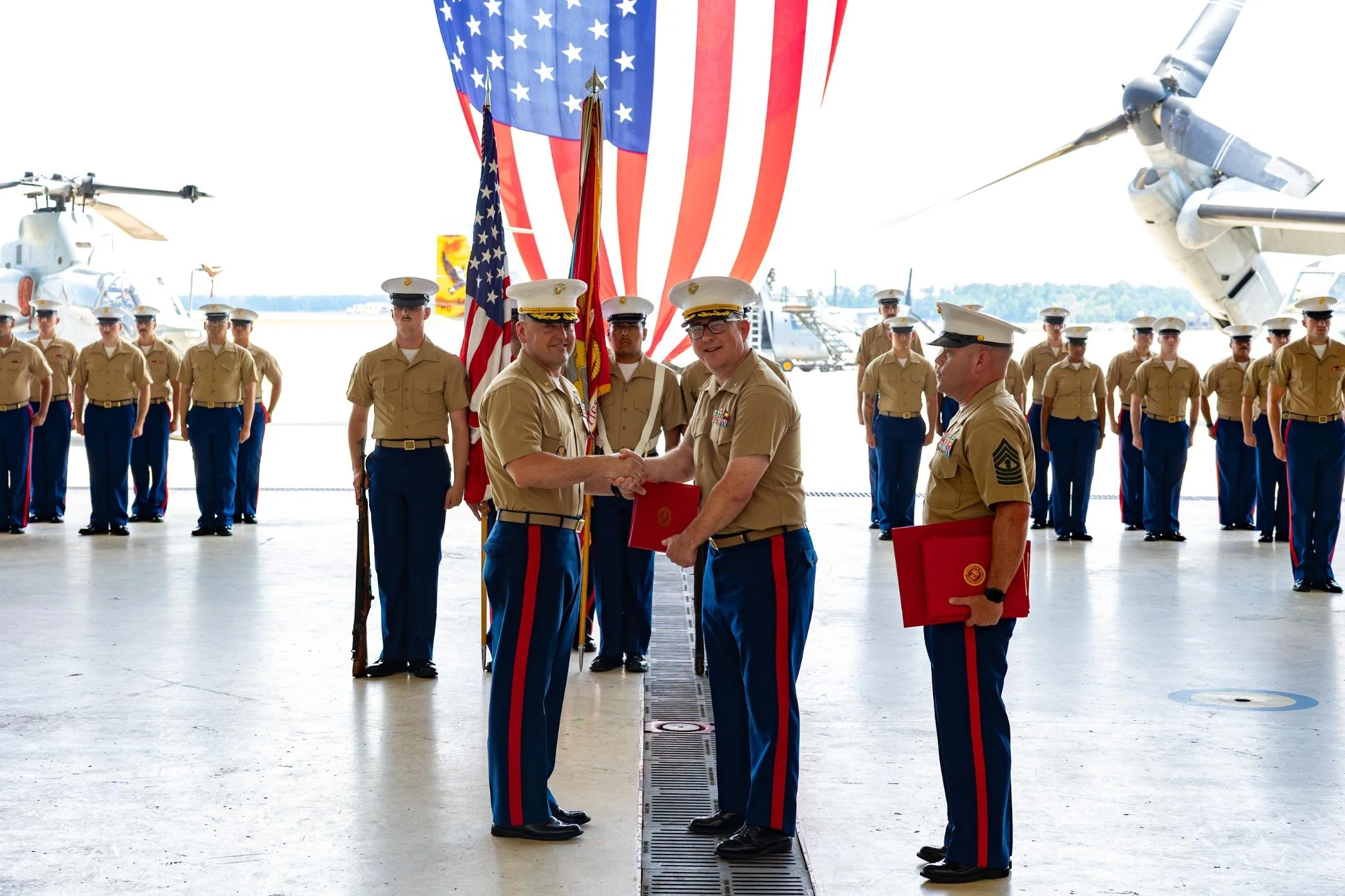Military ceremony at an airbase with soldiers in uniform, an American flag, and military aircraft in the background.