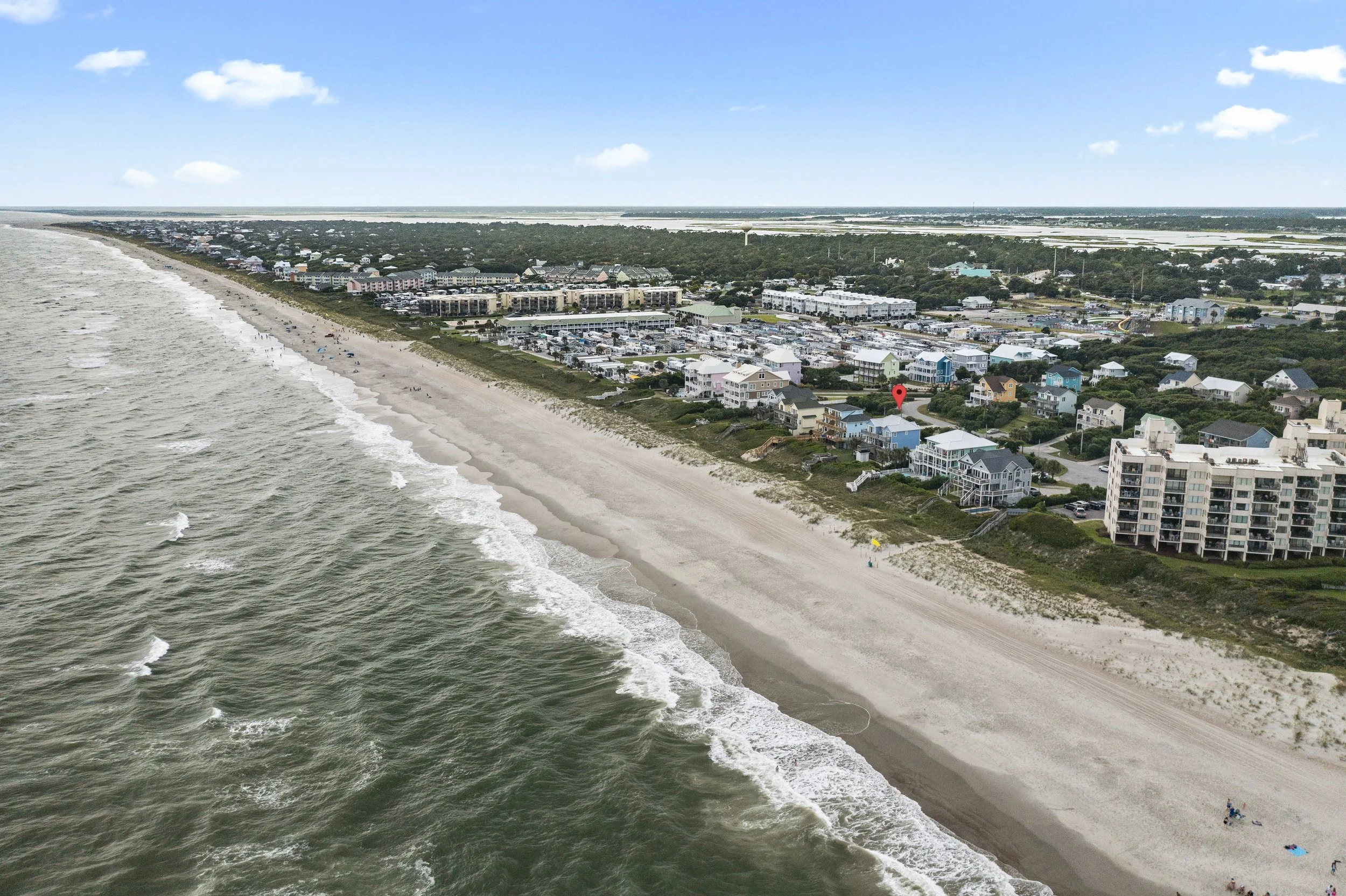 Aerial view of a beach with waves crashing on the shore, and colorful houses and buildings along the coast with a green landscape beyond.