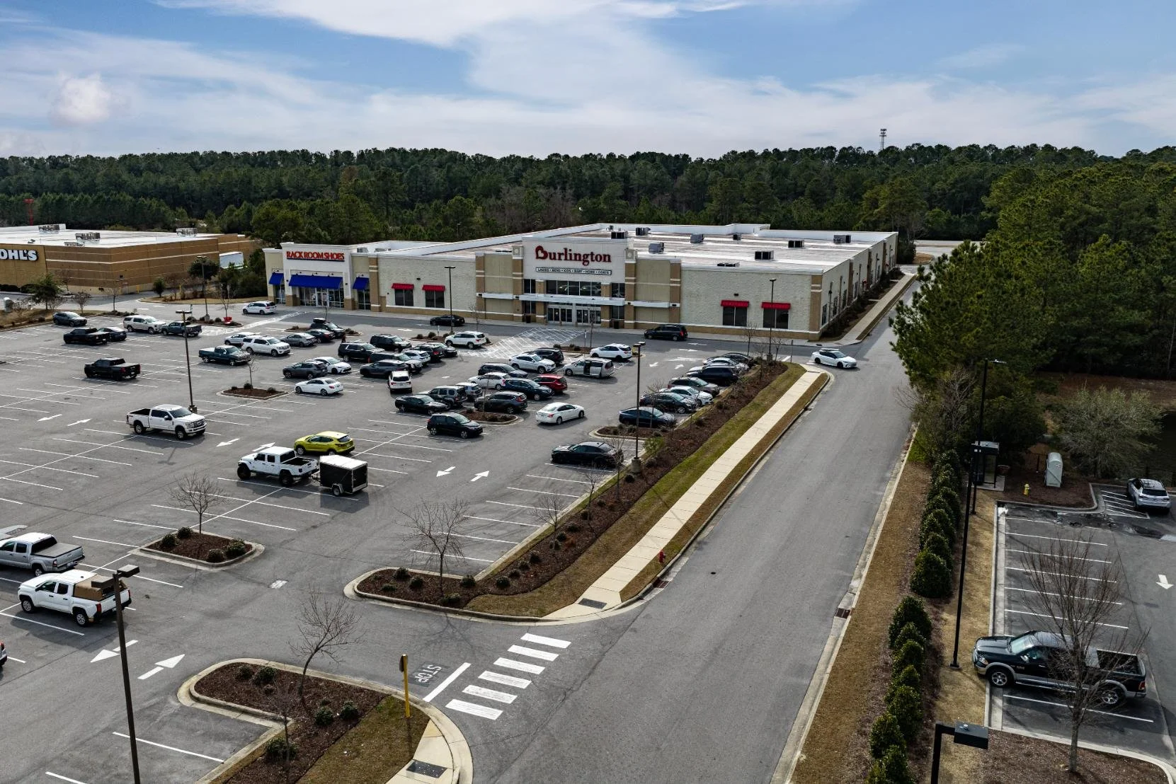 A large shopping mall parking lot with multiple parked cars, a few trees, and a shopping center with stores named Costco, Burlington, and Rack Room Shoes in the background. The parking lot is mostly empty.