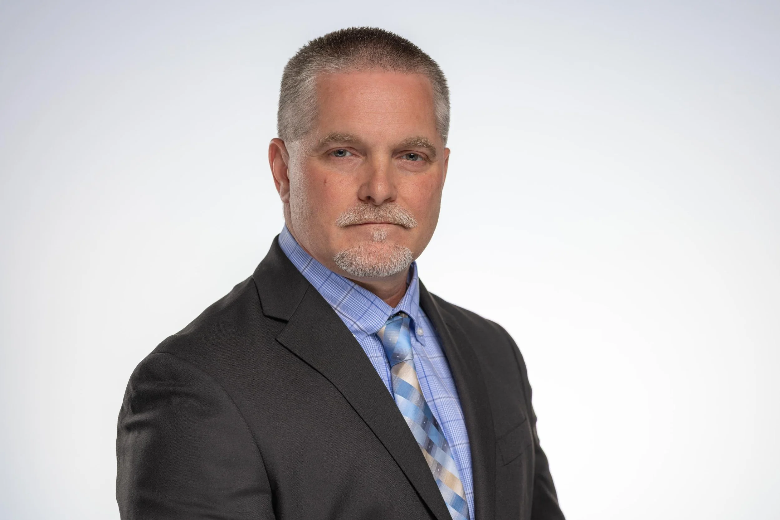 A middle-aged man with short, graying hair and a trimmed beard wearing a dark suit jacket, a blue checkered shirt, and a striped tie, standing against a plain light background.