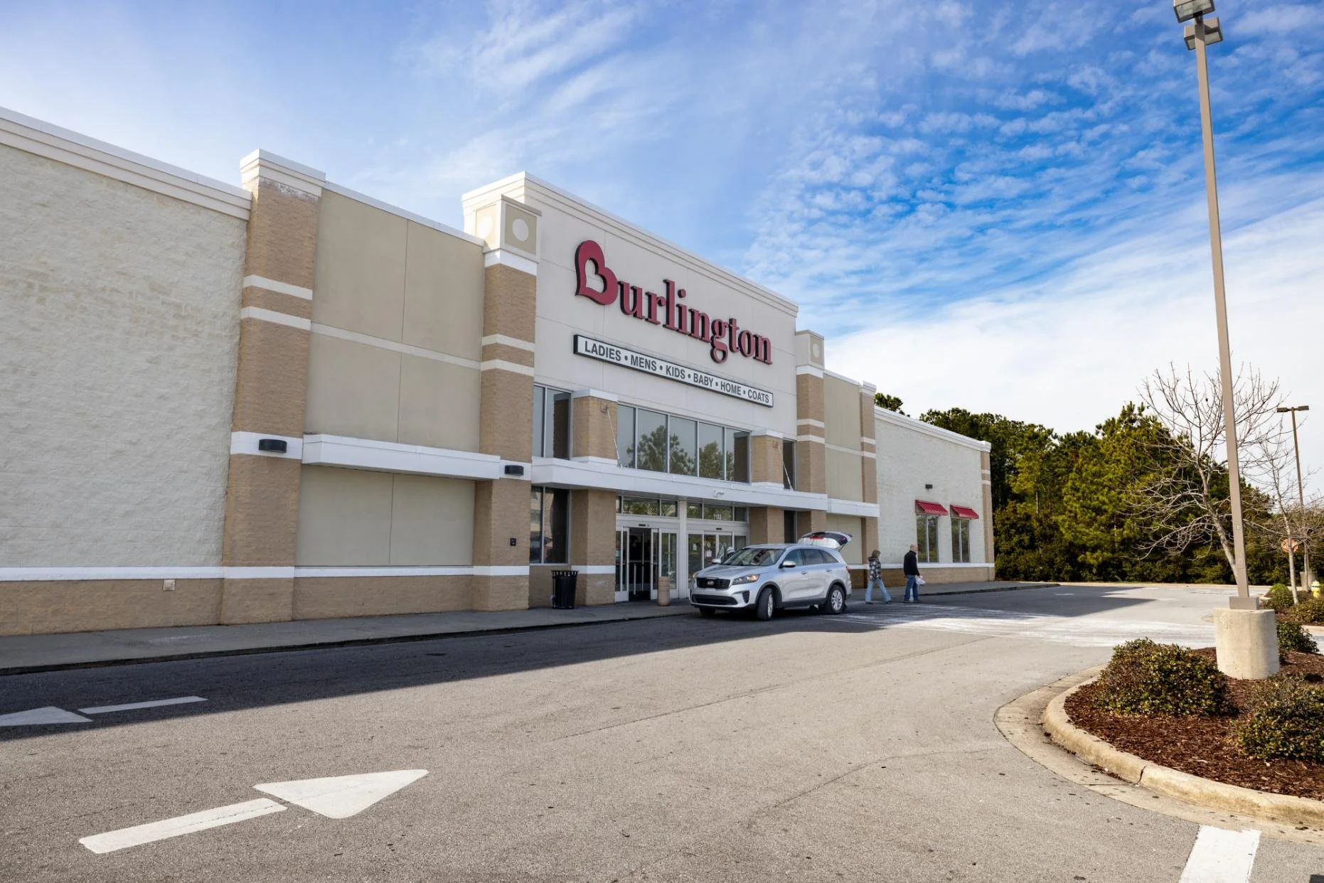 Exterior view of Burlington store with parking lot, a white SUV parked near the entrance, and a few people walking outside, under a partly cloudy sky.