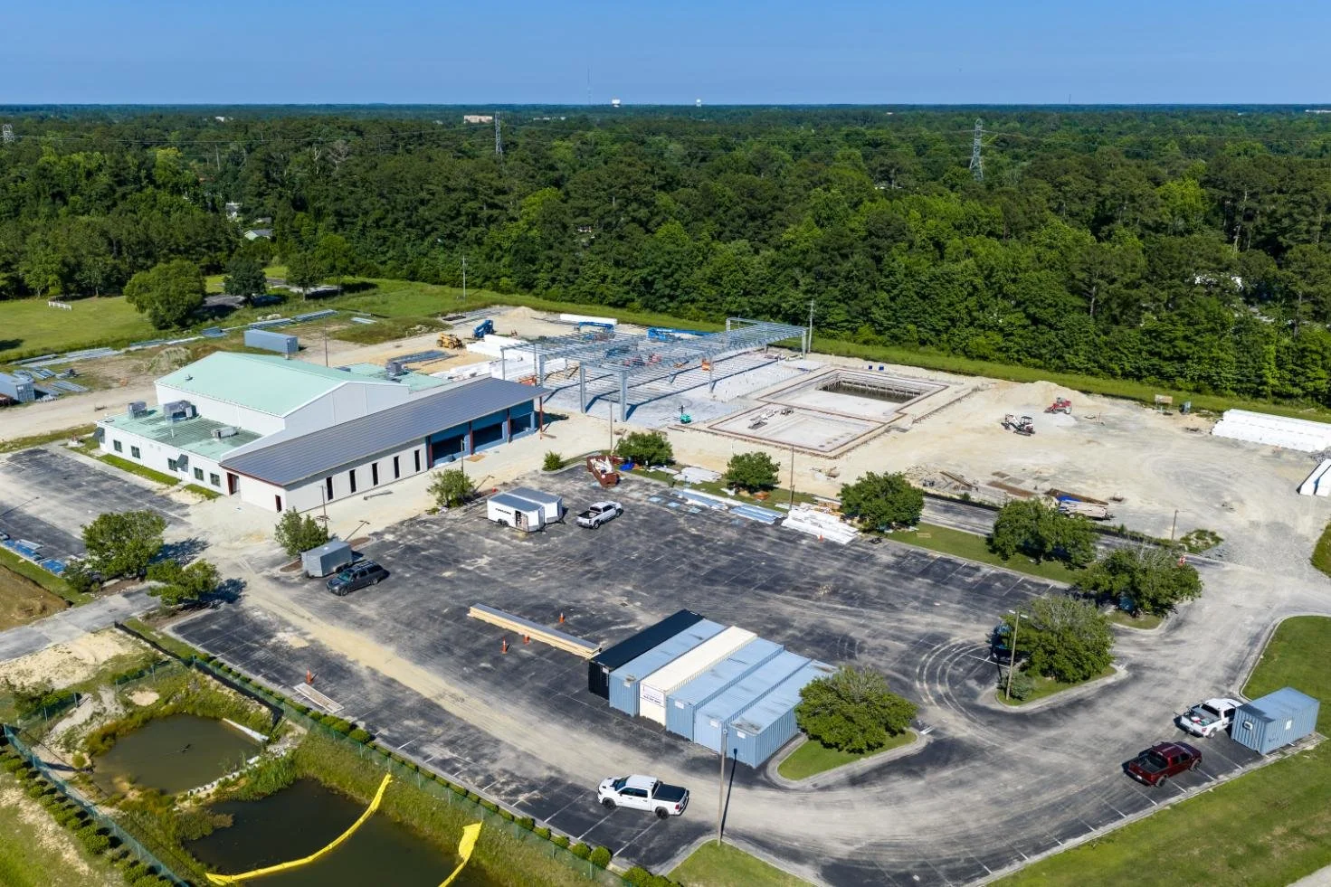 Aerial view of a construction site with a sports complex under development, including a large building, parking lot, and a pond, surrounded by green trees and open land.