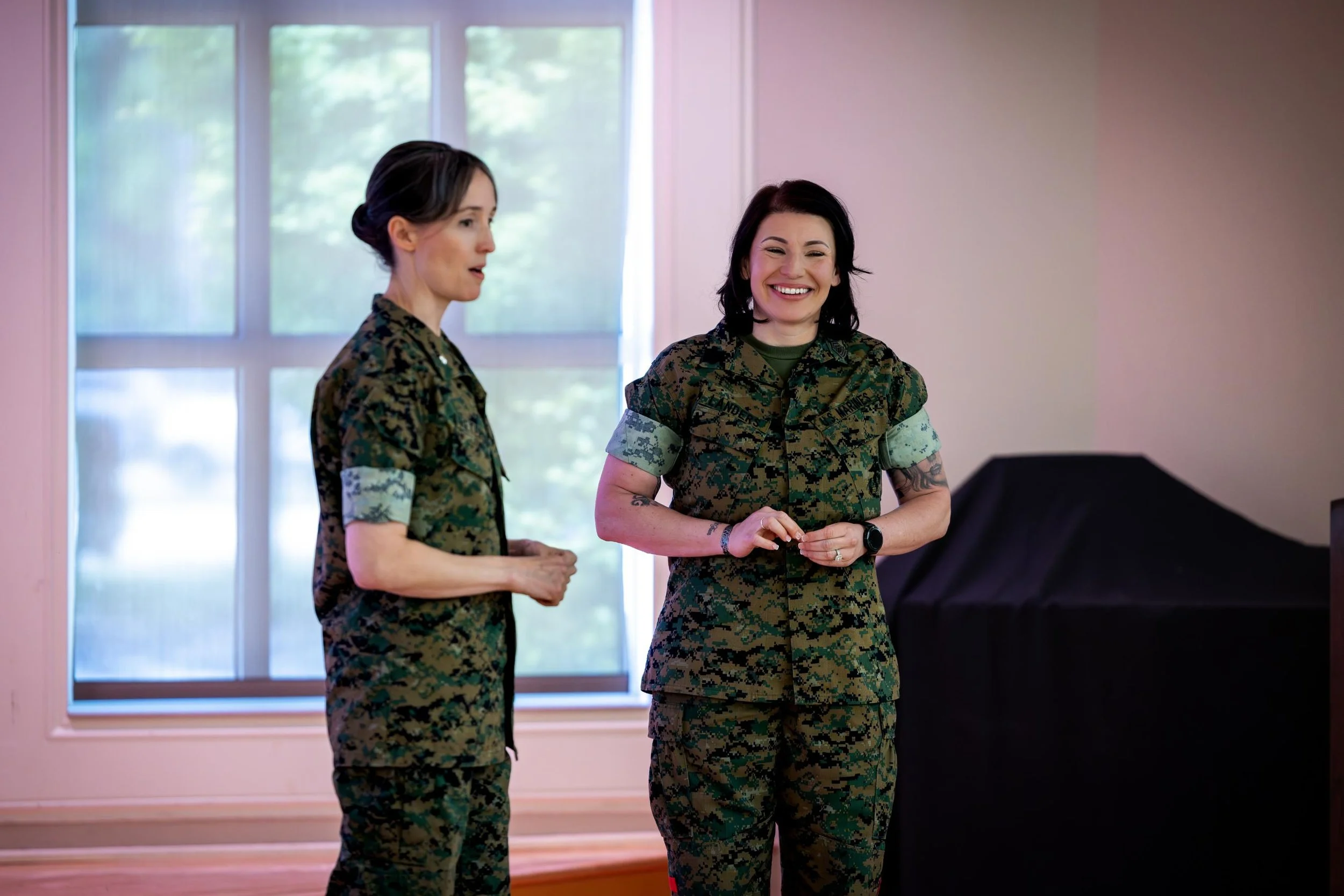 Two women in camouflage military uniforms standing and talking inside a room with large windows.