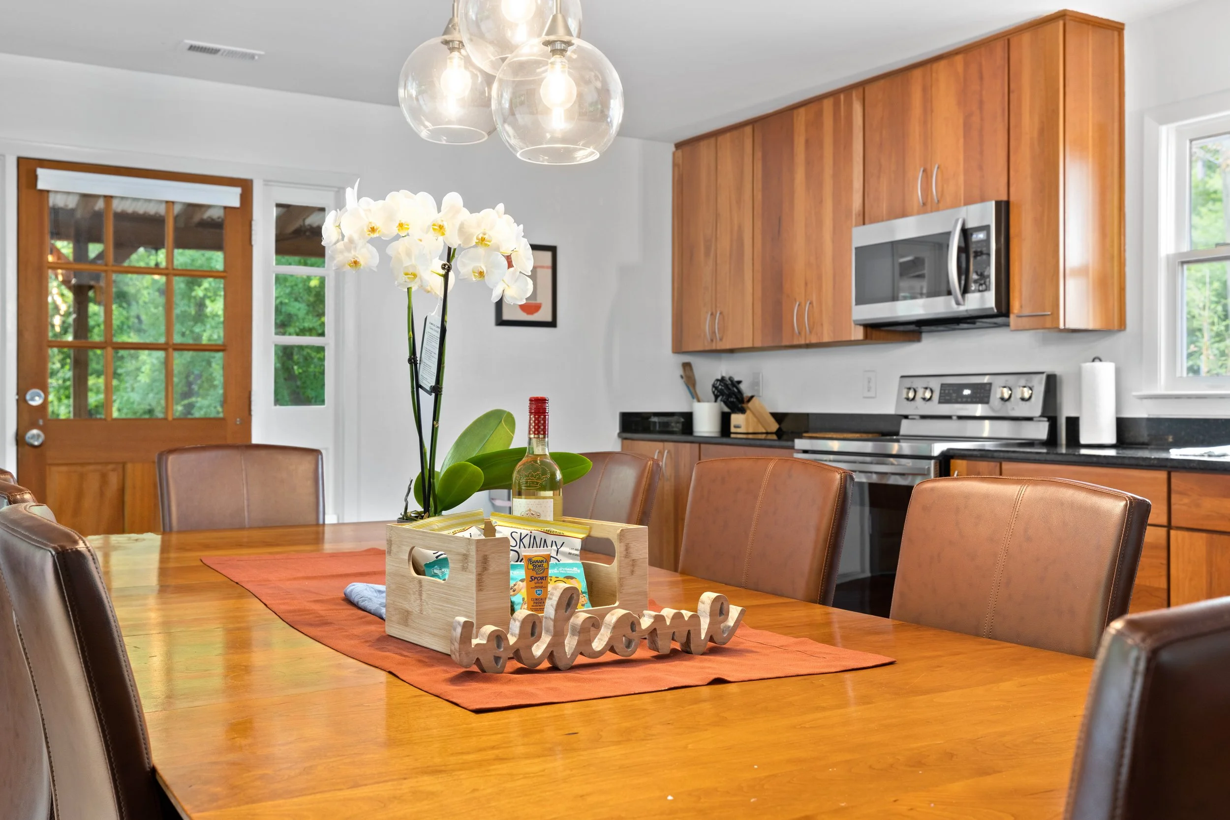 A wooden dining table with brown leather chairs, a vase with white orchids, a bottle of wine, and a decorative wooden sign that spells 'welcome'. In the background, there is a kitchen with wooden cabinets, stainless steel appliances, and a window.