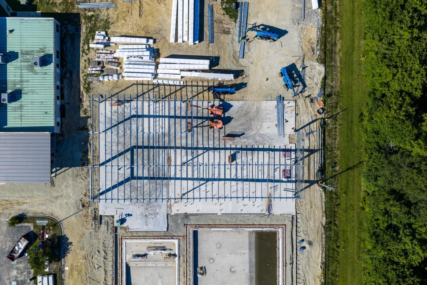 Aerial view of a construction site with a steel framework for a building, construction equipment, stacks of building materials, and surrounding greenery.