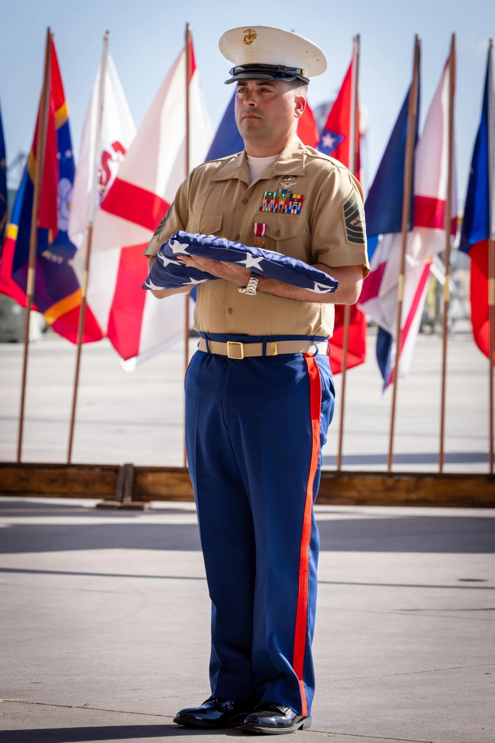 A United States Marine in dress uniform holding a folded American flag during a formal ceremony with American flags in the background.